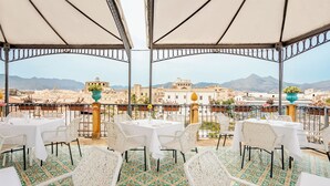Un área de comedor al aire libre con mesas y sillas blancas, piso con diseño y vista a edificios y montañas al fondo.