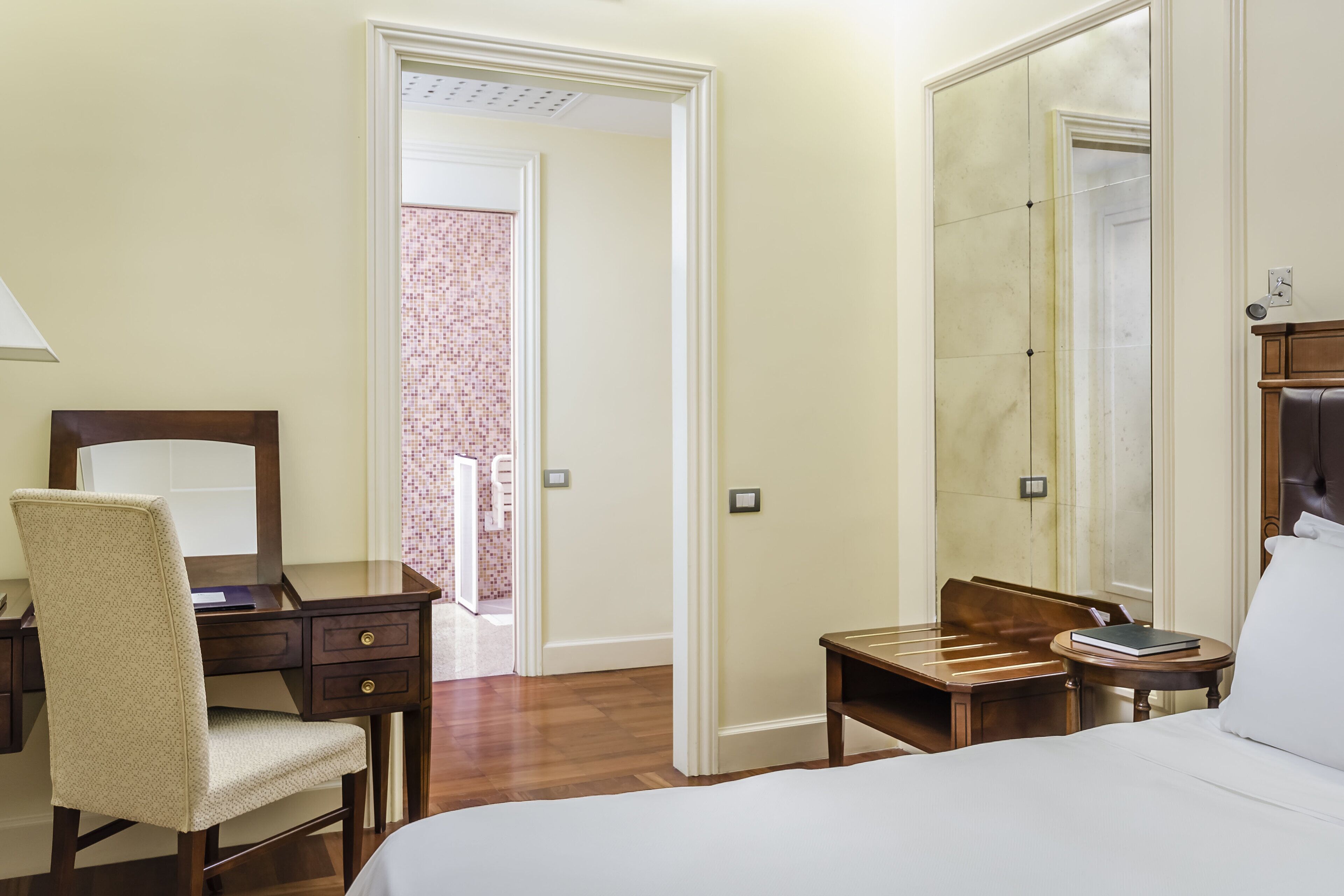 Habitación de hotel con cama, escritorio con silla, una mesita con un libro y un espejo grande.