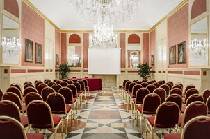 Una sala conferenze con file di sedie rosse, uno schermo bianco grande e un lampadario.