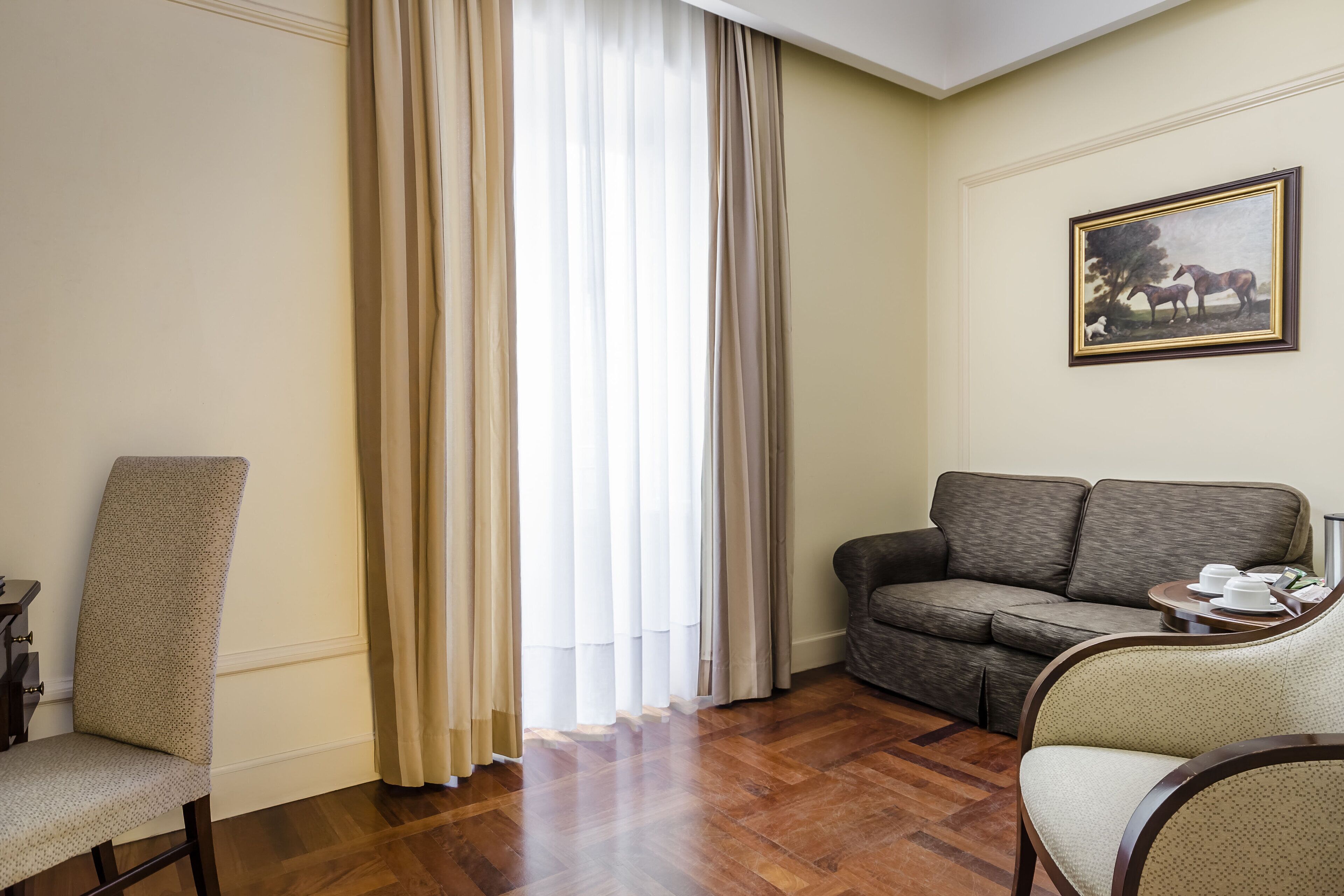 A hotel room with a wooden floor, a grey sofa, a beige armchair, and a painting of horses on the wall.