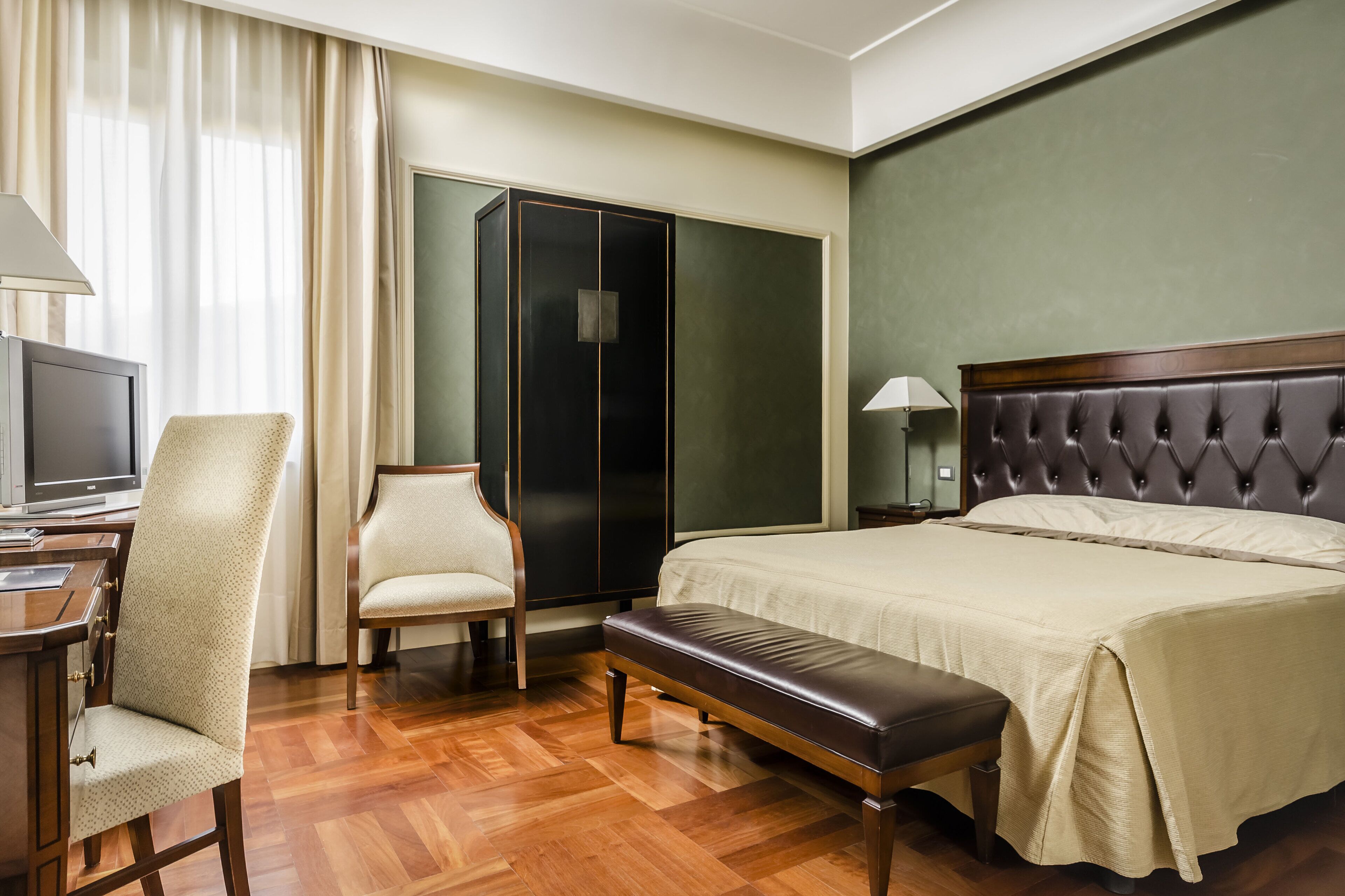 A hotel room with a bed, a desk, a chair, a bench, a wardrobe, and a television.