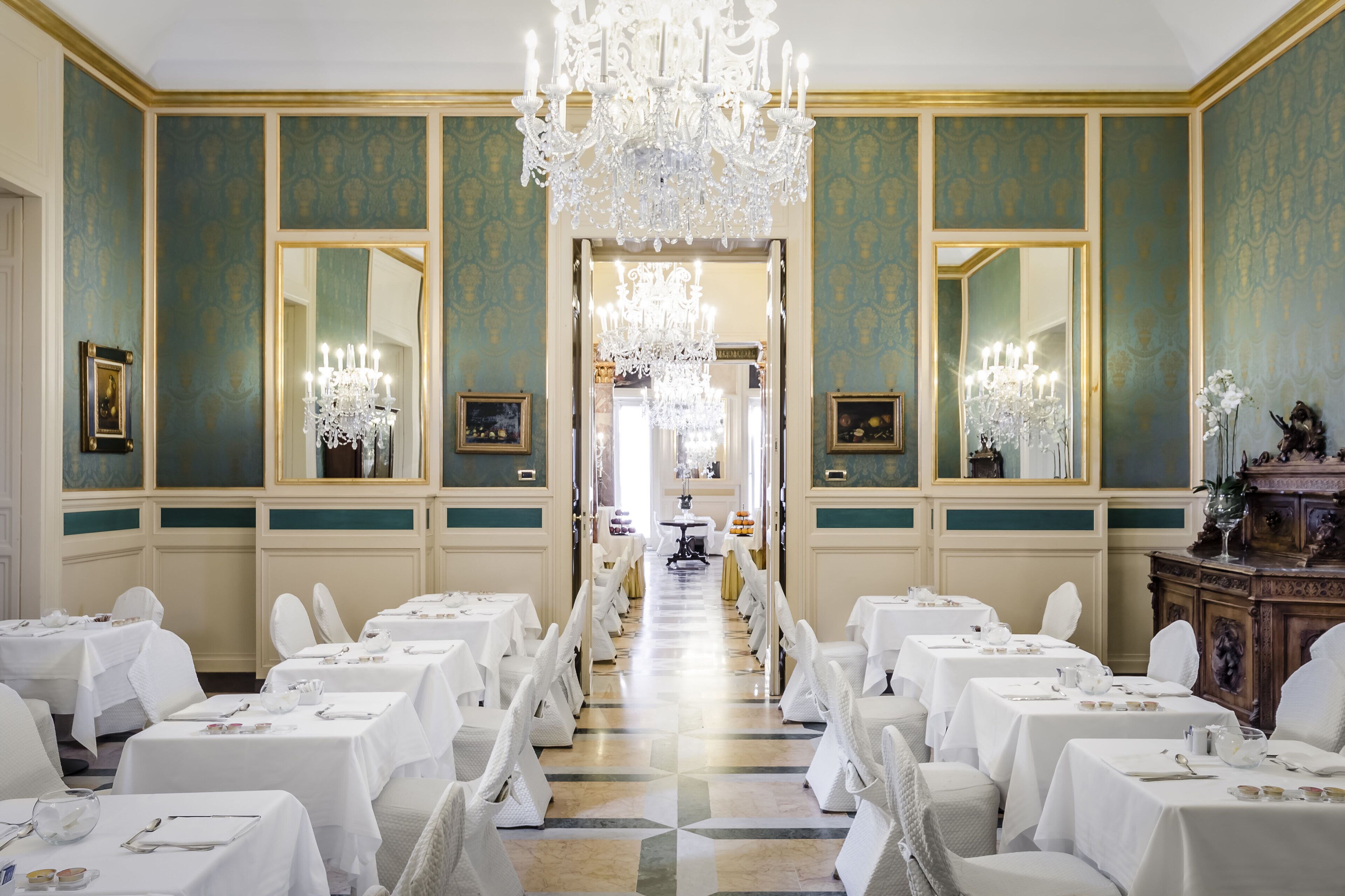 Une salle à manger élégante, avec plusieurs tables dressées pour le service, un grand lustre et des miroirs richement décorés.