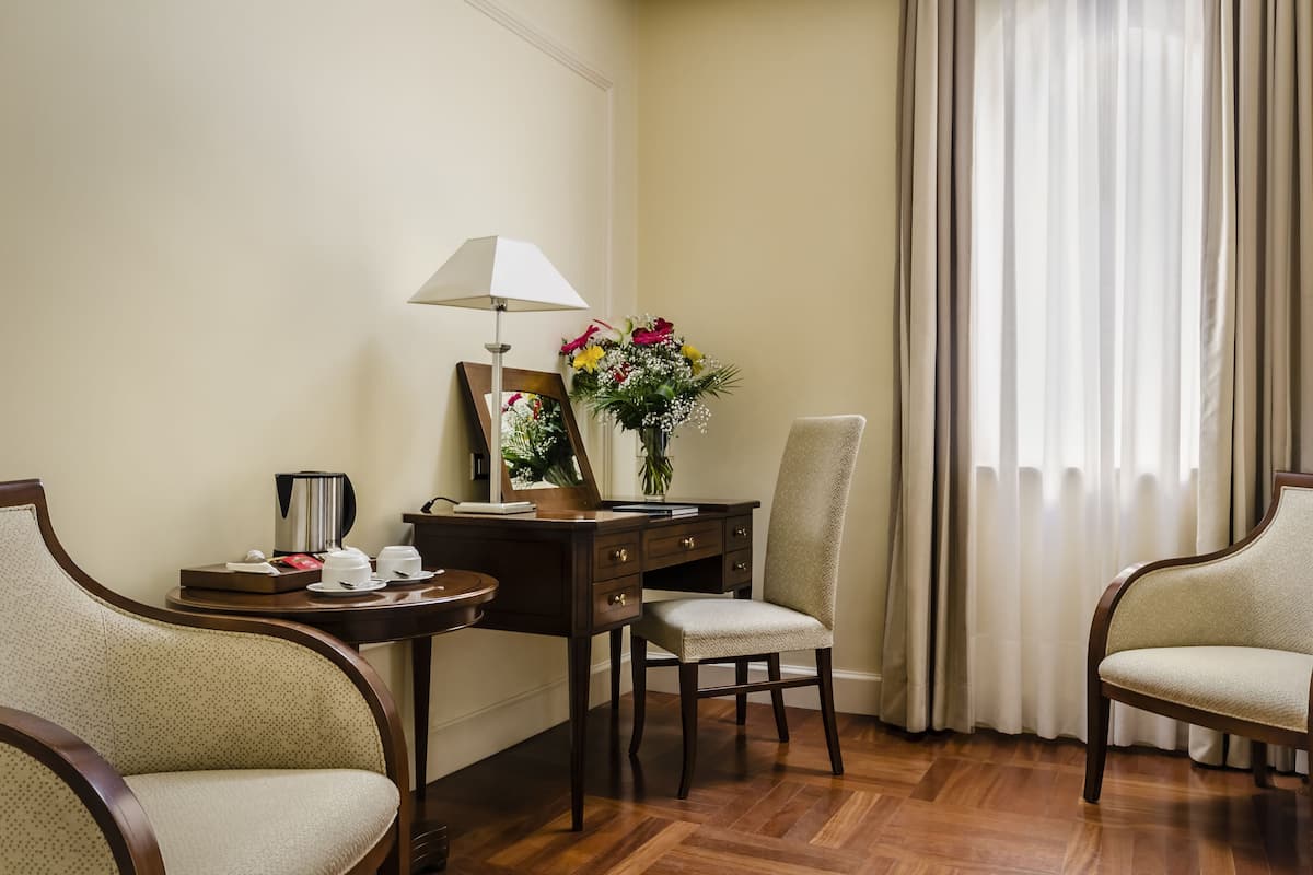 A hotel room with a wooden desk, two armchairs, a lamp, a mirror, a vase with flowers, and a view of the outside through sheer curtains.