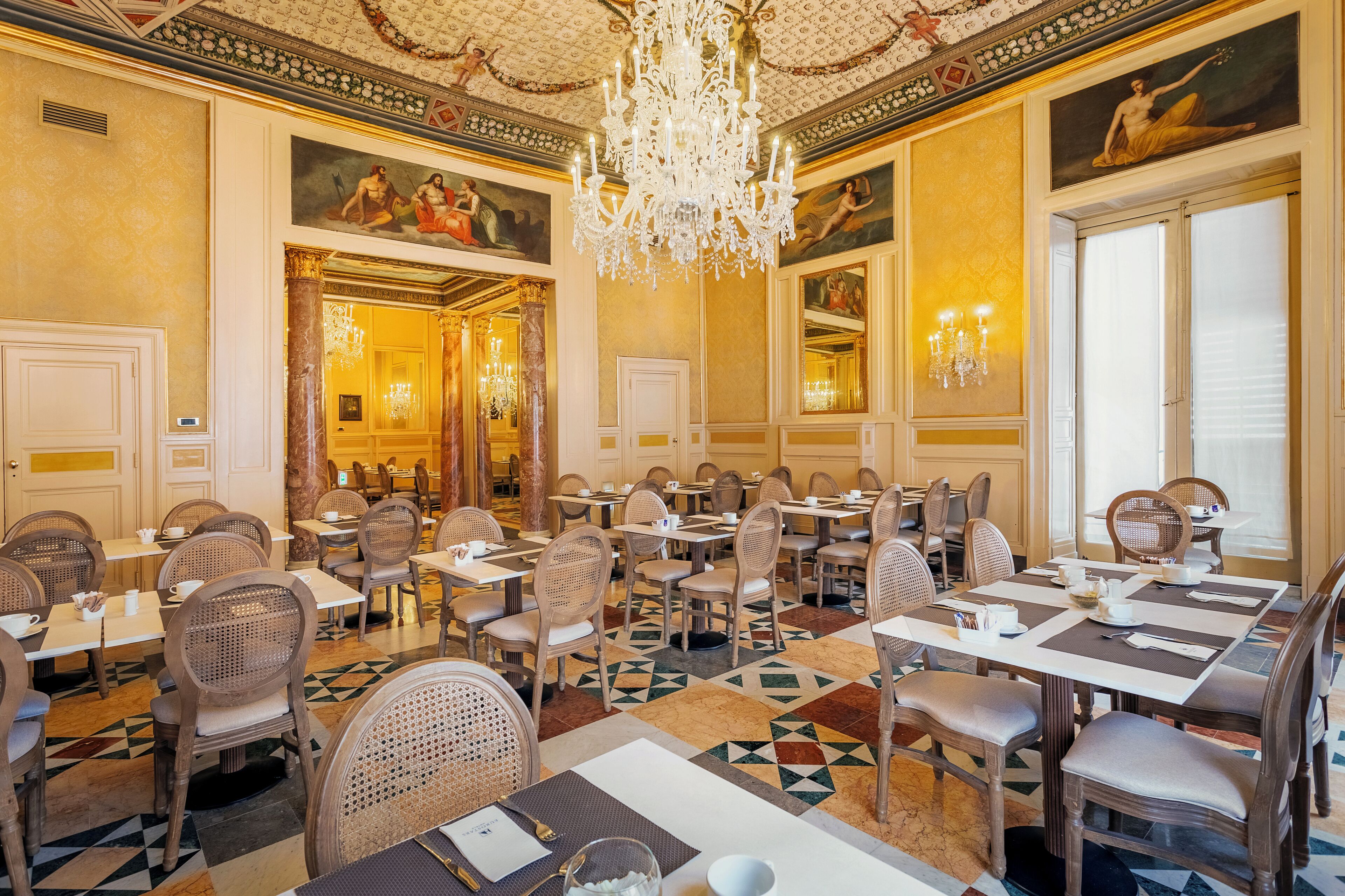 A formal dining room with multiple tables and chairs, a chandelier, and classical wall paintings.