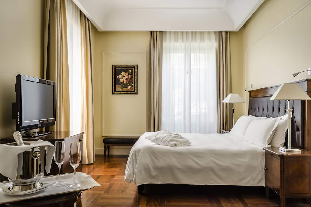 A hotel room with a bed, bedside lamps, a television, a minibar, and a window with curtains.