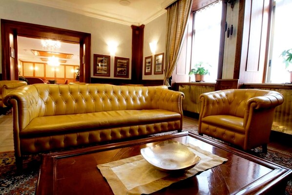 Una habitación con un sofá grande y capitonado y un sillón a juego, una mesa de centro de madera con un cuenco decorativo, cuadros en la pared y un ventanal con cortinas.