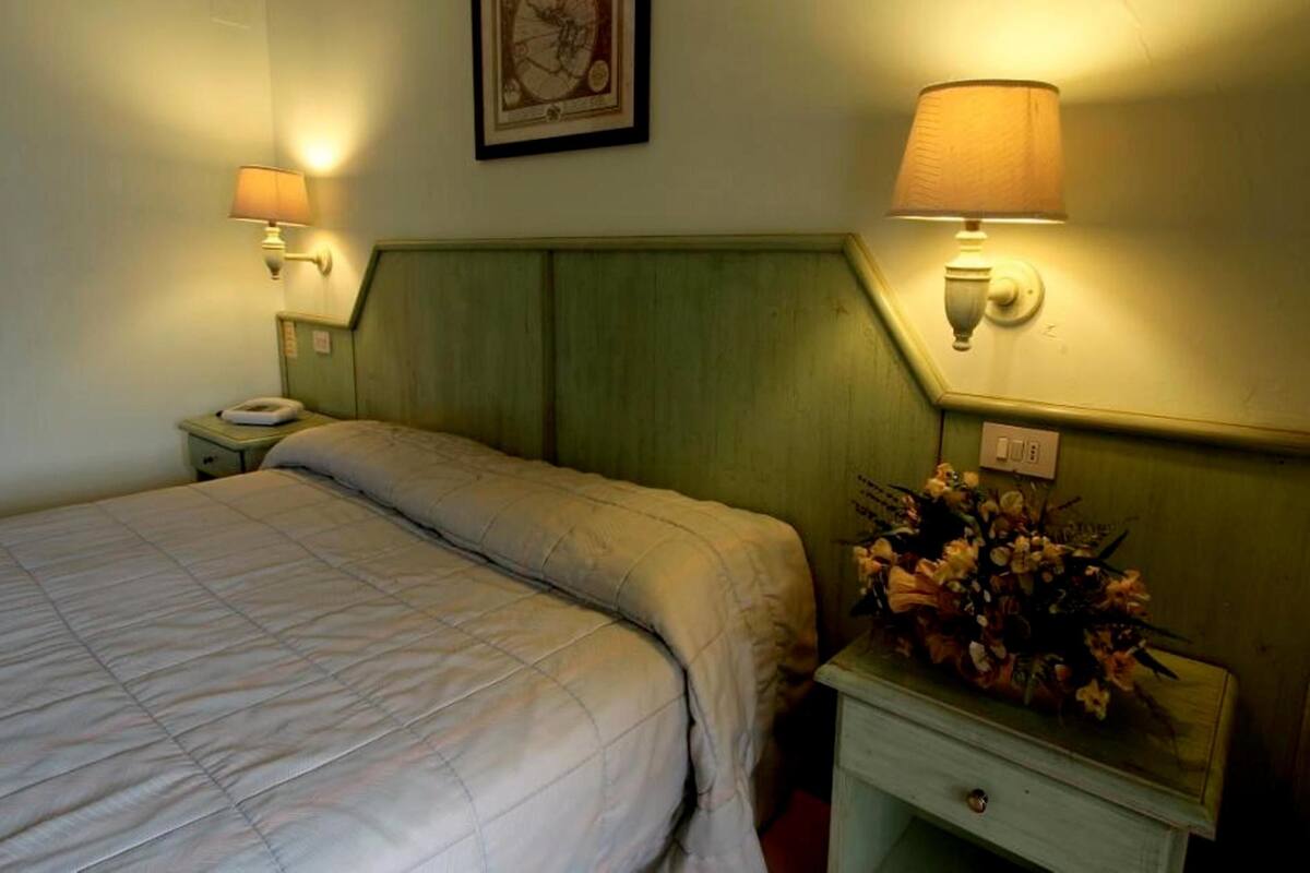 A hotel room with a bed, two bedside lamps, a nightstand with a phone, and a wall-mounted light switch.