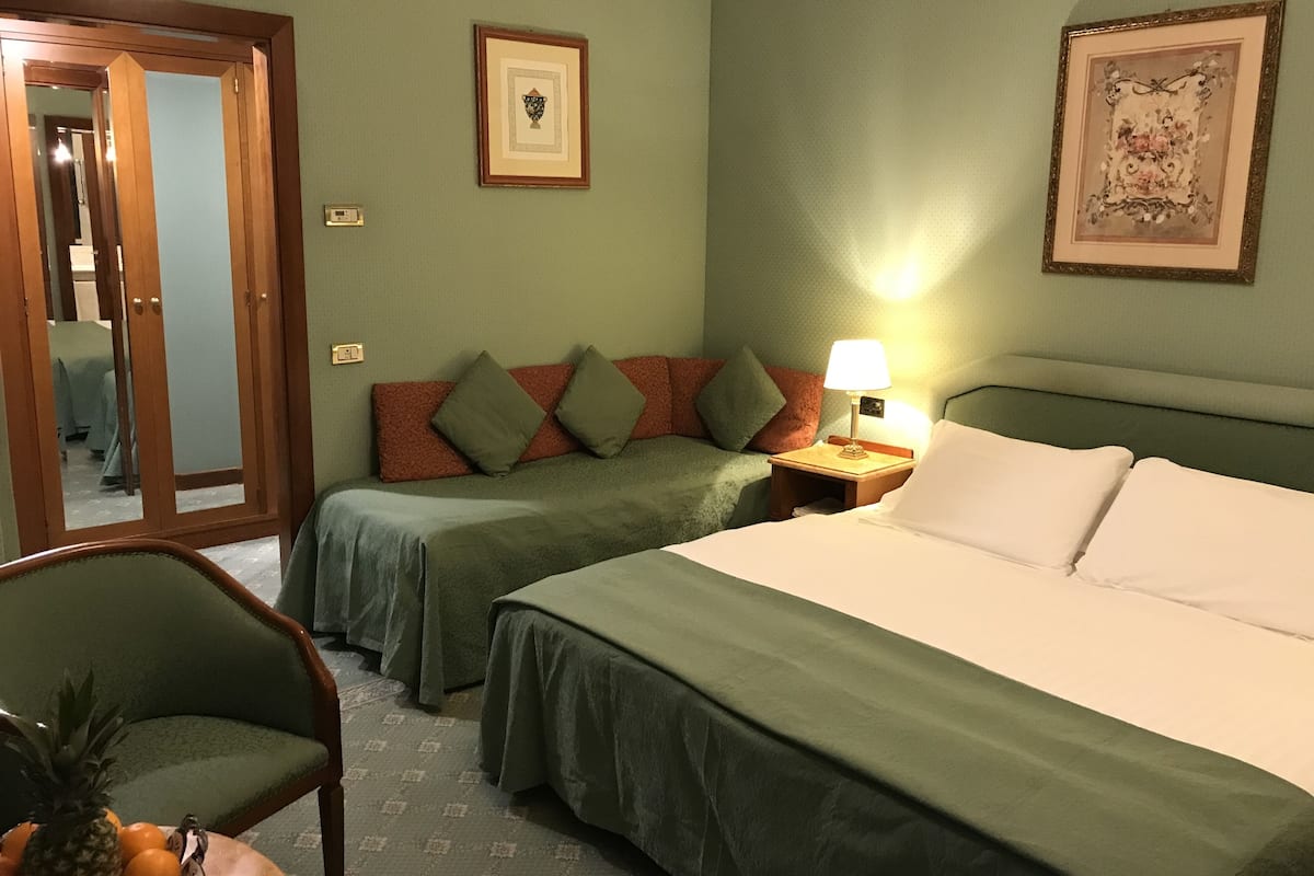 A hotel room with two beds, a nightstand with a lamp, a chair, a fruit bowl, and a door leading to another room.