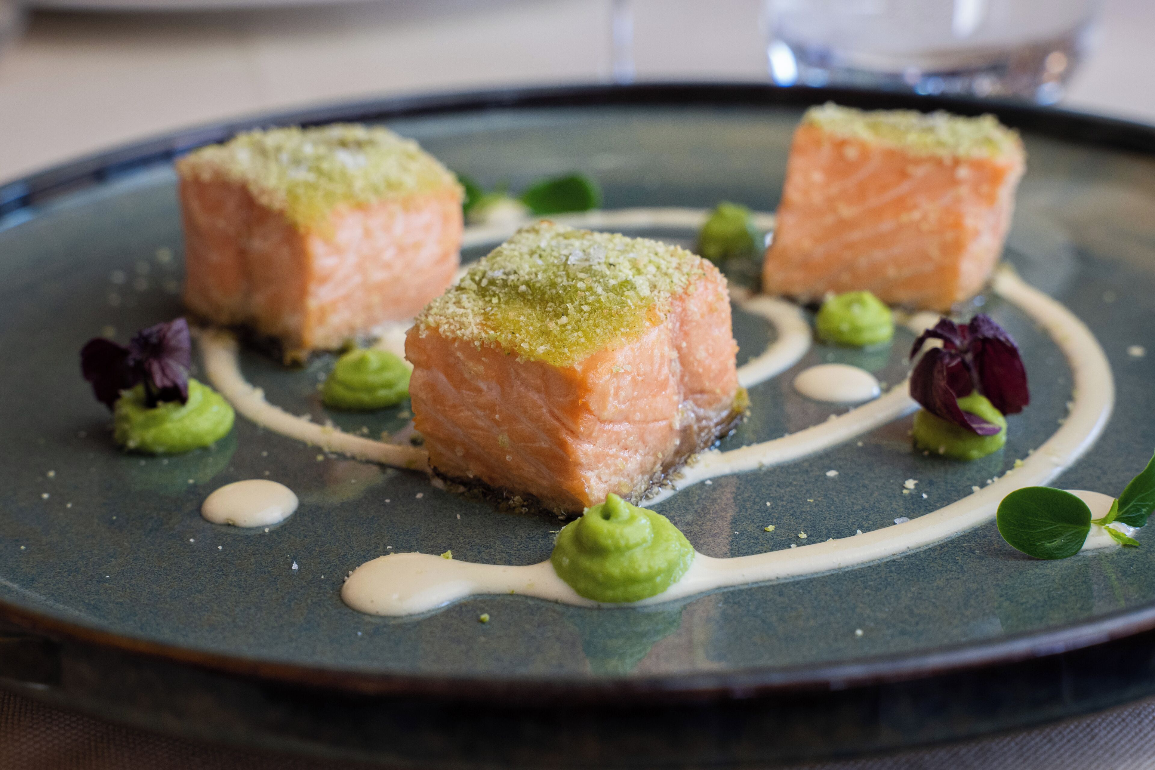 Ein Teller mit drei Lachsfilets, belegt mit grüner Dekoration, umgeben von kleinen Klecksen grüner Soße und violetten Blüten.
