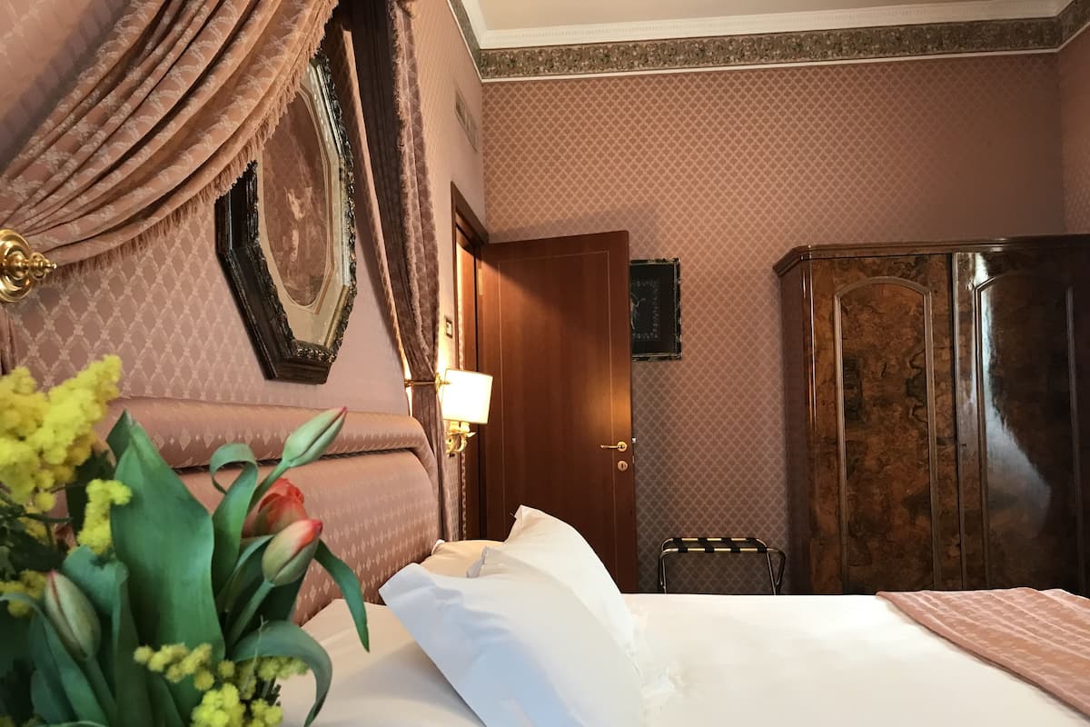 A hotel room with a large bed, a wooden wardrobe, a nightstand with a lamp, and a decorative mirror on the wall.
