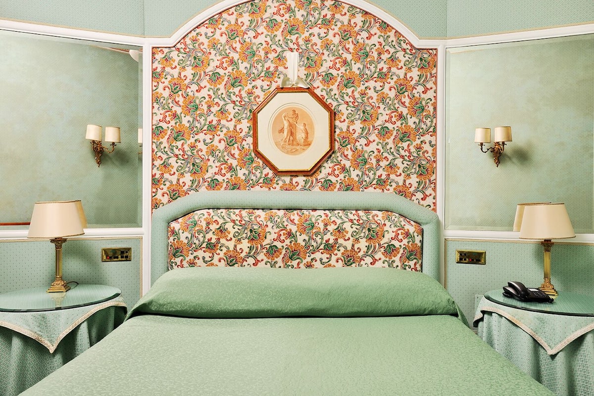 A room with a floral wallpaper, a bed with a green bedspread, two bedside tables with lamps, and a framed picture on the wall.