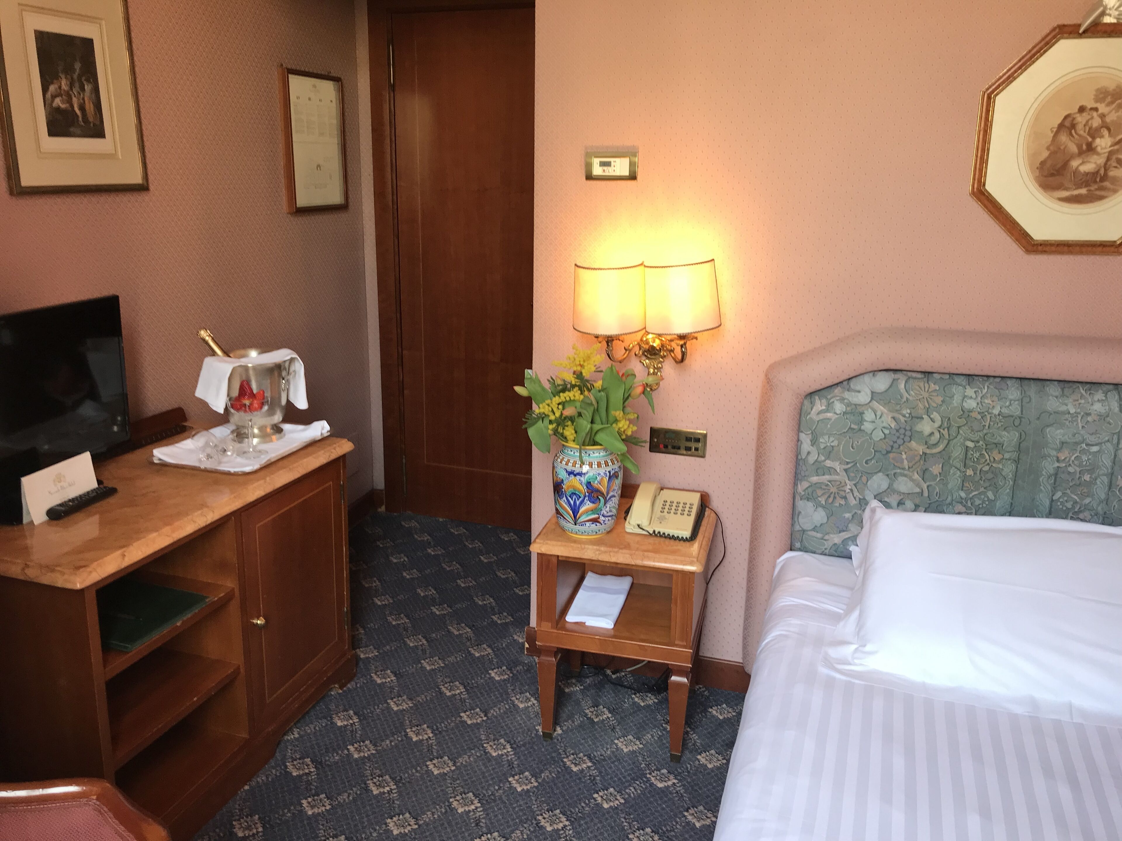 A hotel room with a bed, a desk with a computer, a lamp, a vase with flowers, and a telephone.