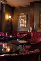 A dimly lit room with a patterned wallpaper, a large framed picture on the wall, and several red upholstered chairs and tables.