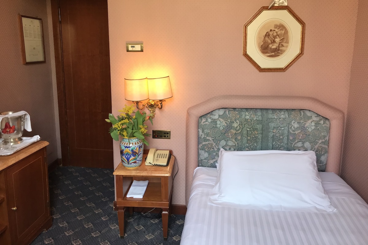 A hotel room with a bed, a bedside table with a lamp, a vase with flowers, a telephone, and a framed picture on the wall.