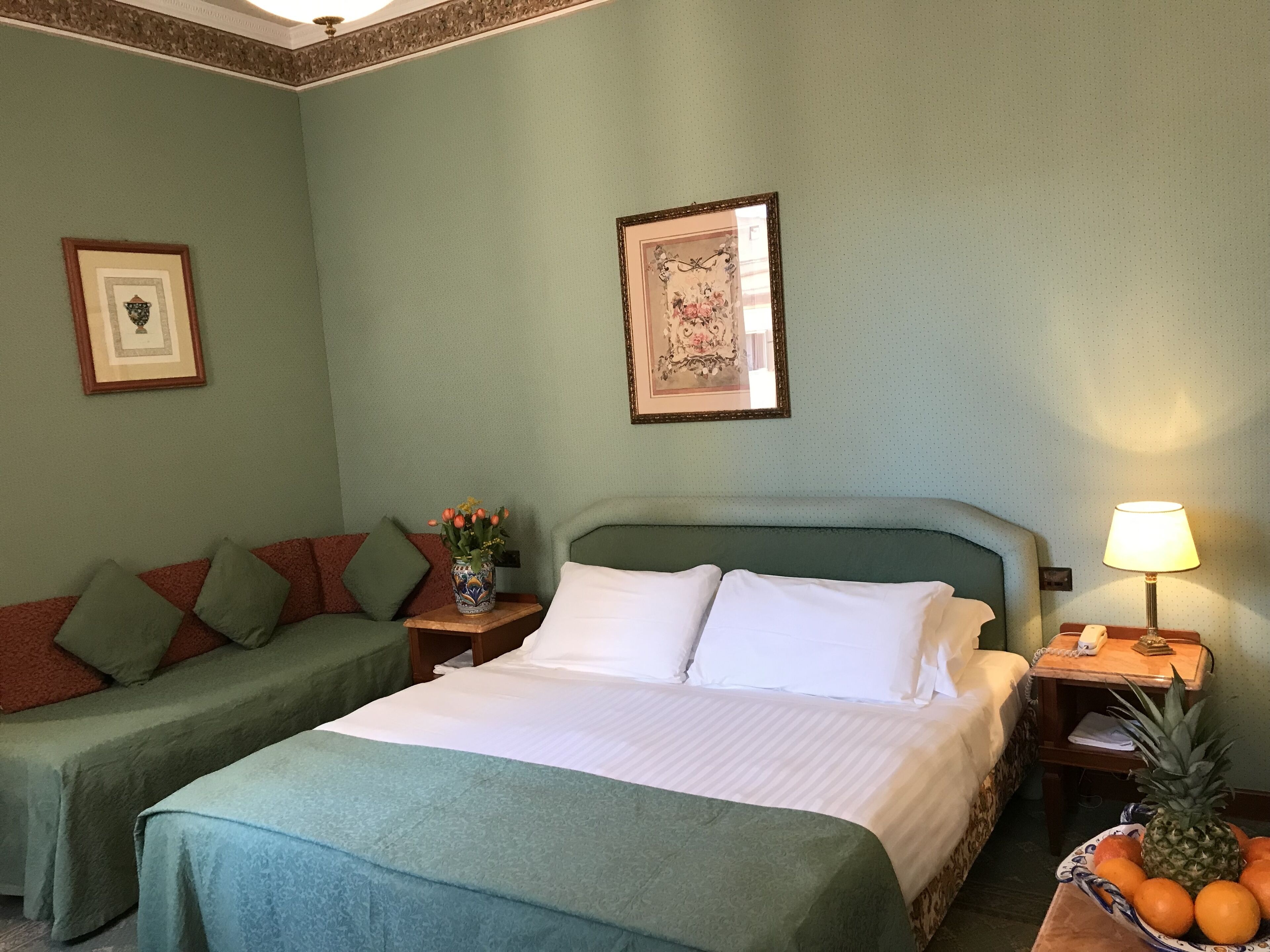 A hotel room with two single beds, a green sofa, a vase with flowers, a framed picture, a lamp, and a fruit bowl.