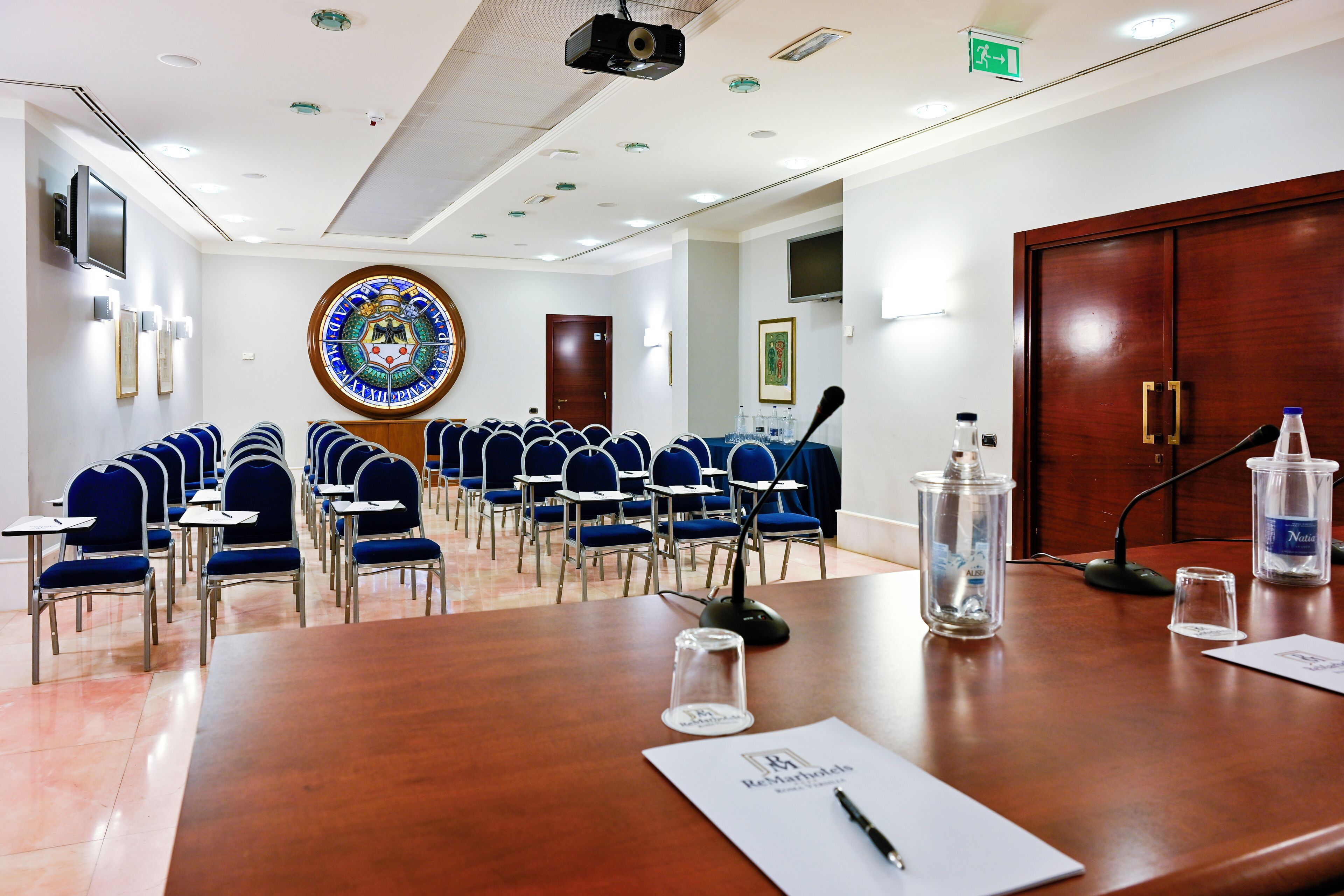 Uma sala de conferências com uma grande mesa de madeira, fileiras de cadeiras azuis, um projetor e um amplo vitral.