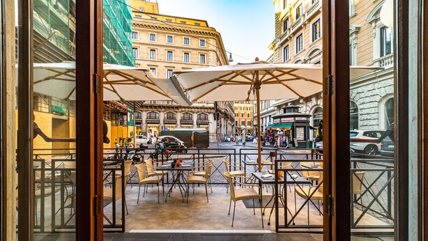 Un café al aire libre con mesas y sillas, vista a la calle y edificios al fondo.