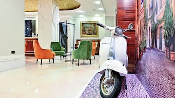 Un vestíbulo con un scooter blanco, sillas verdes y anaranjadas, un suelo de mármol y un mural de una escena callejera.