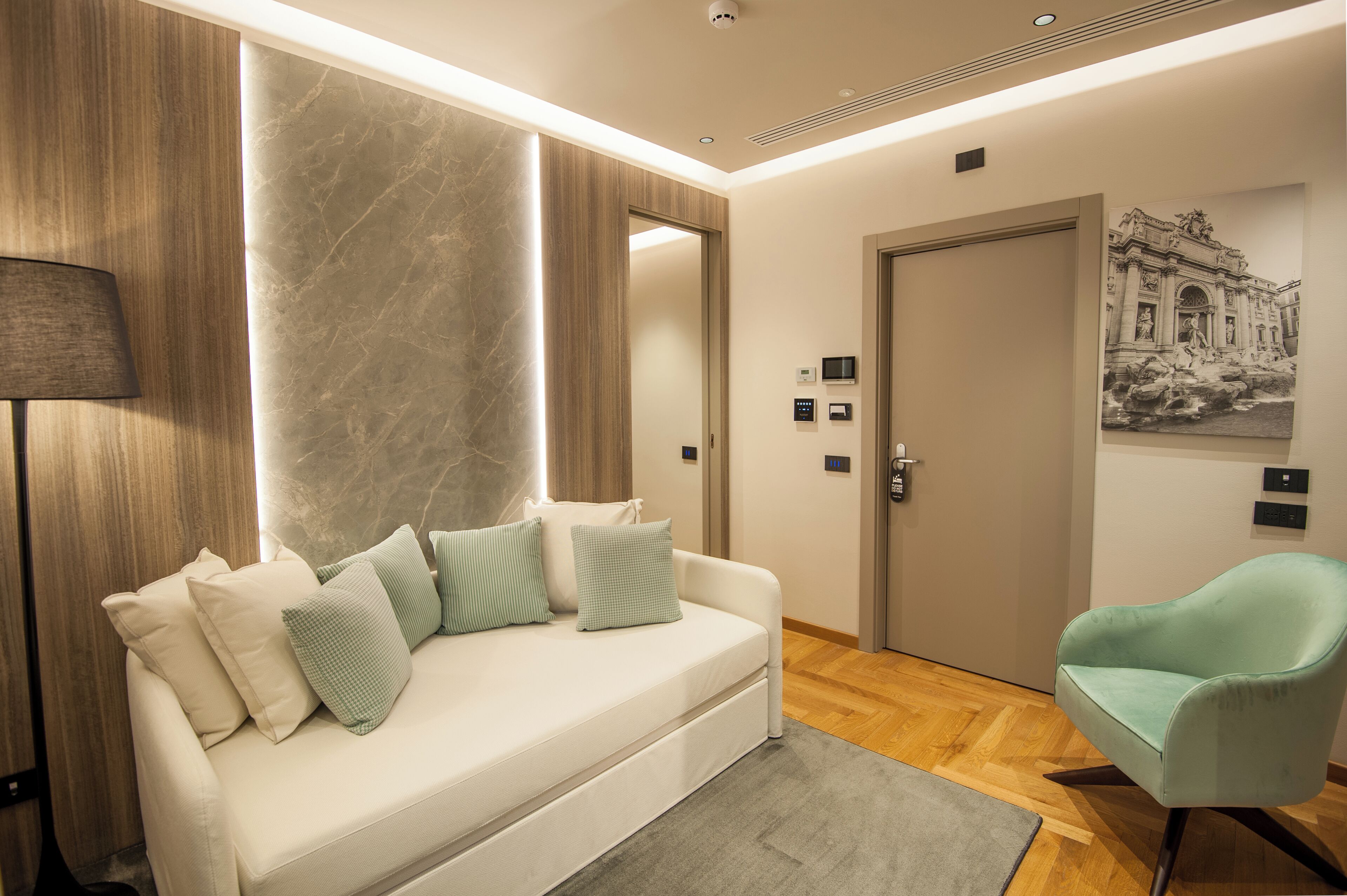 A modern hotel room with a white sofa, teal armchair, and a marble accent wall.
