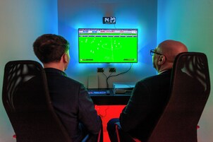 Two people seated facing a large screen displaying a soccer game, with a digital clock above it.