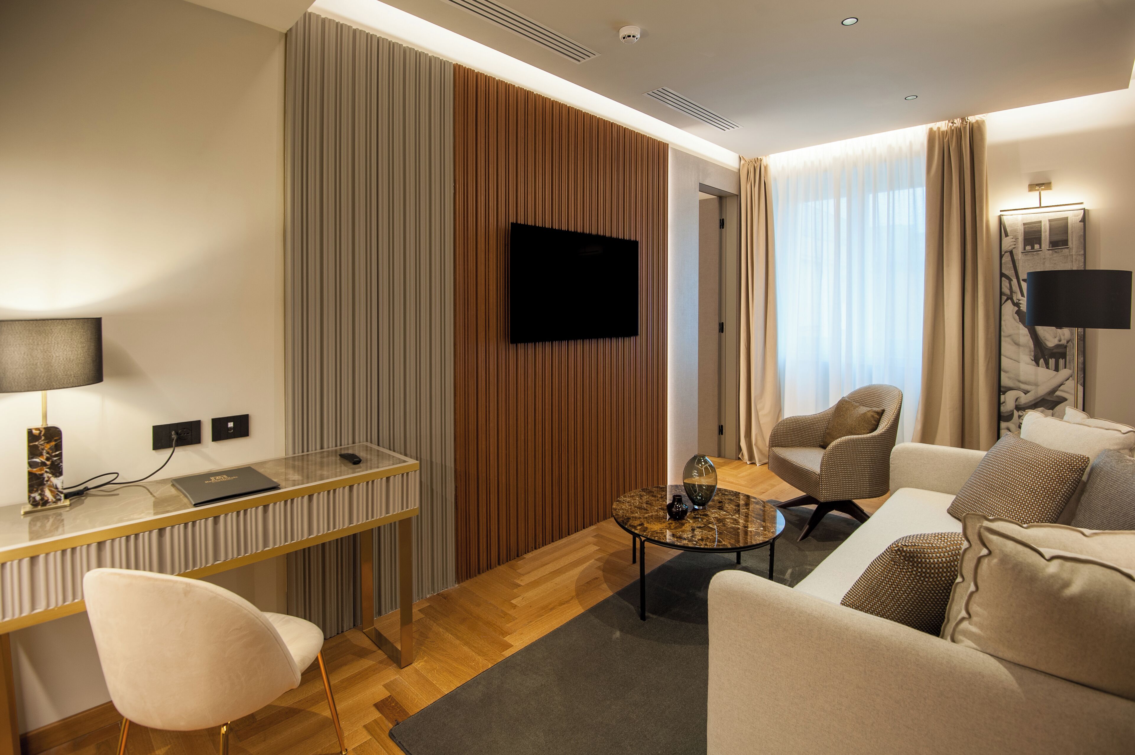 A modern hotel room with a flat-screen TV mounted on the wall, a dining area with a glass table, and a seating area with a sofa and armchairs.