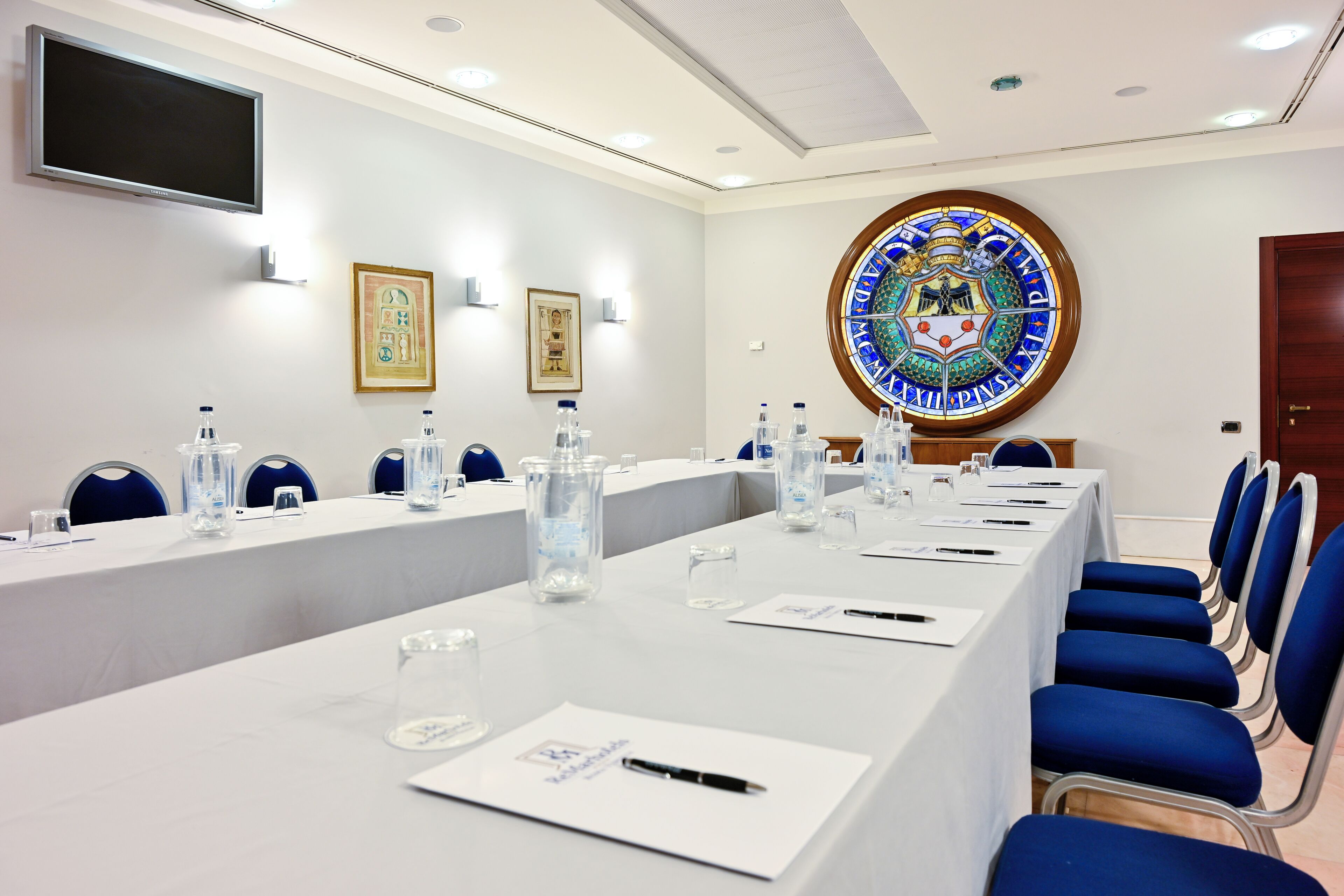 Ein Konferenzraum mit einem langen Tisch, Stühlen, Wasserflaschen, Namensschildern und einem großen Emblem an der Wand.