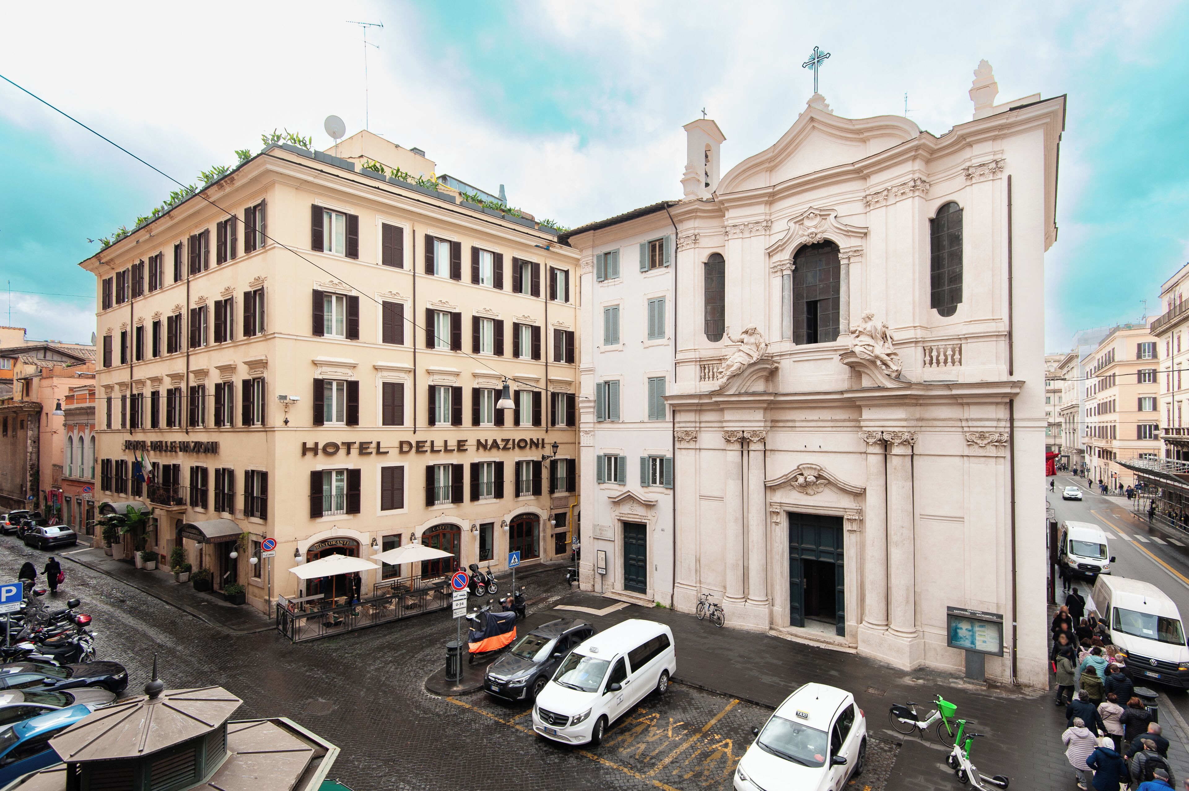 A hotel with a classic facade, a central entrance, and a street view with parked cars and a crowd.