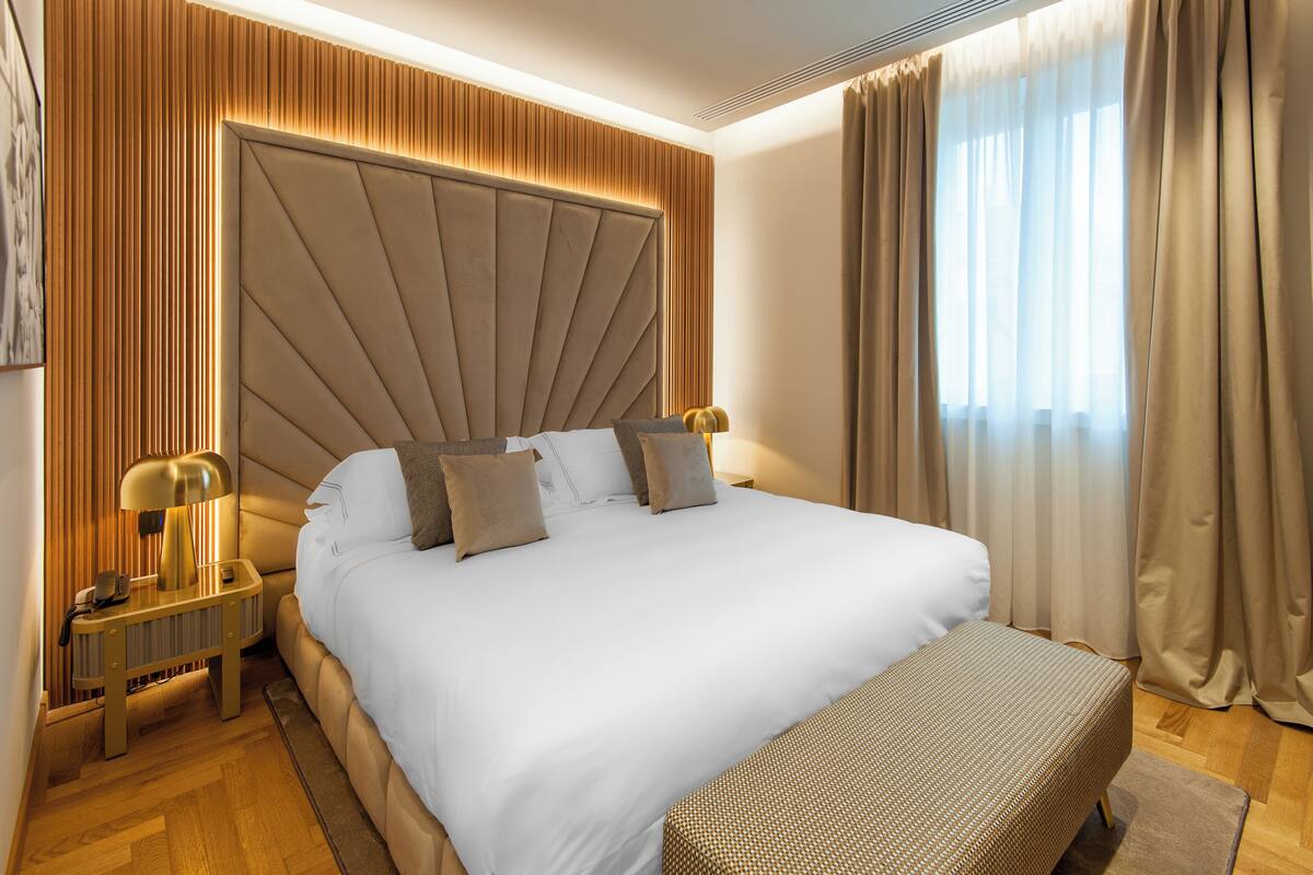 Habitación de hotel moderna con una cama grande, una mesita de noche con lámpara y vistas al exterior a través de cortinas translúcidas.
