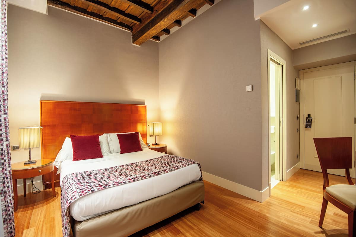 Un dormitorio ordenado con cabecera de madera, una cama grande con colcha estampada, dos lámparas de noche y una silla de madera.