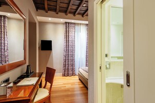 A hotel room with a wooden desk, chair, and a bed. There is a television mounted on the wall and a window with curtains.