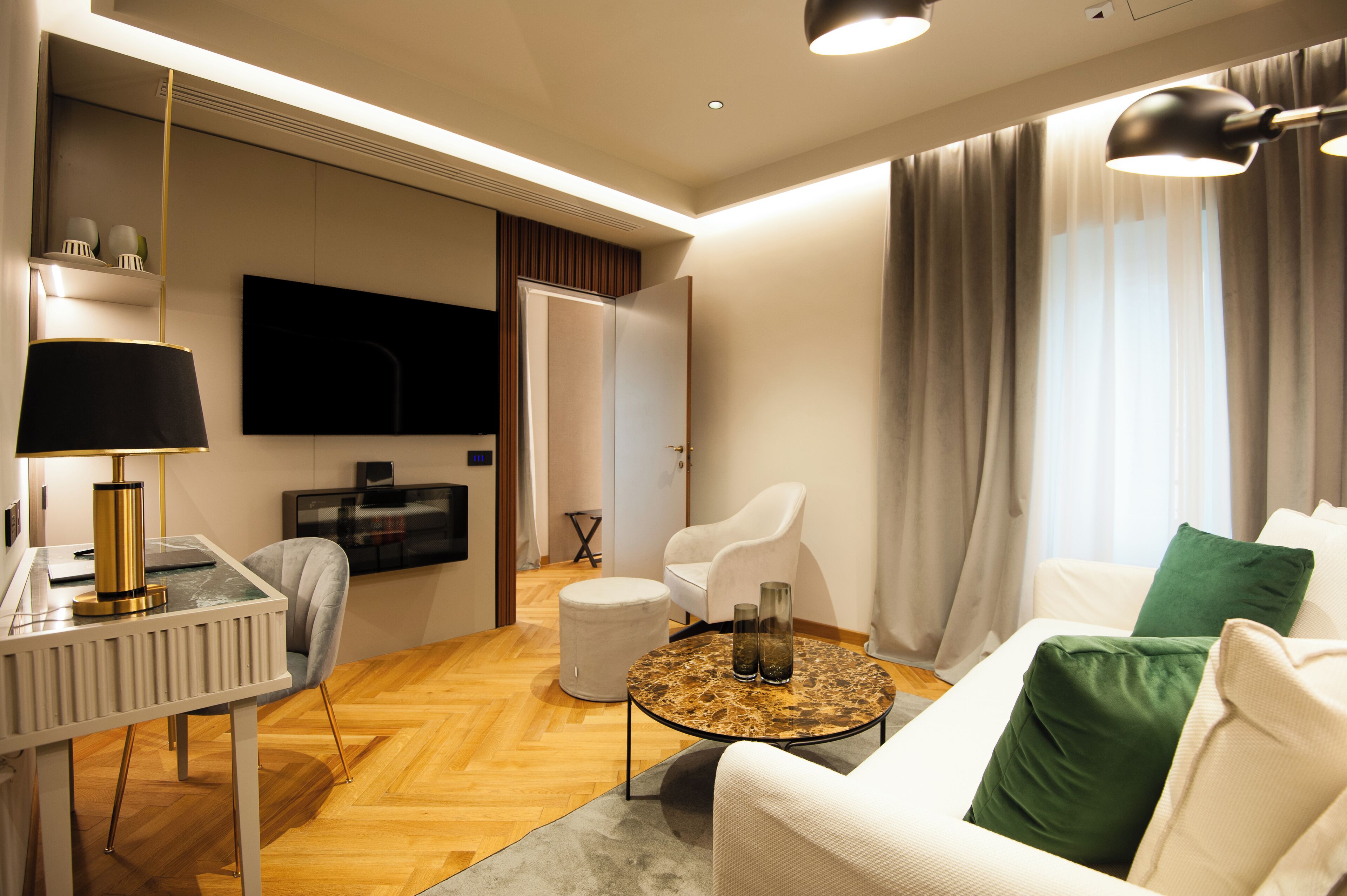 A modern living room with a white sofa, a round coffee table, a white armchair, a TV mounted on the wall, and a large window with curtains.