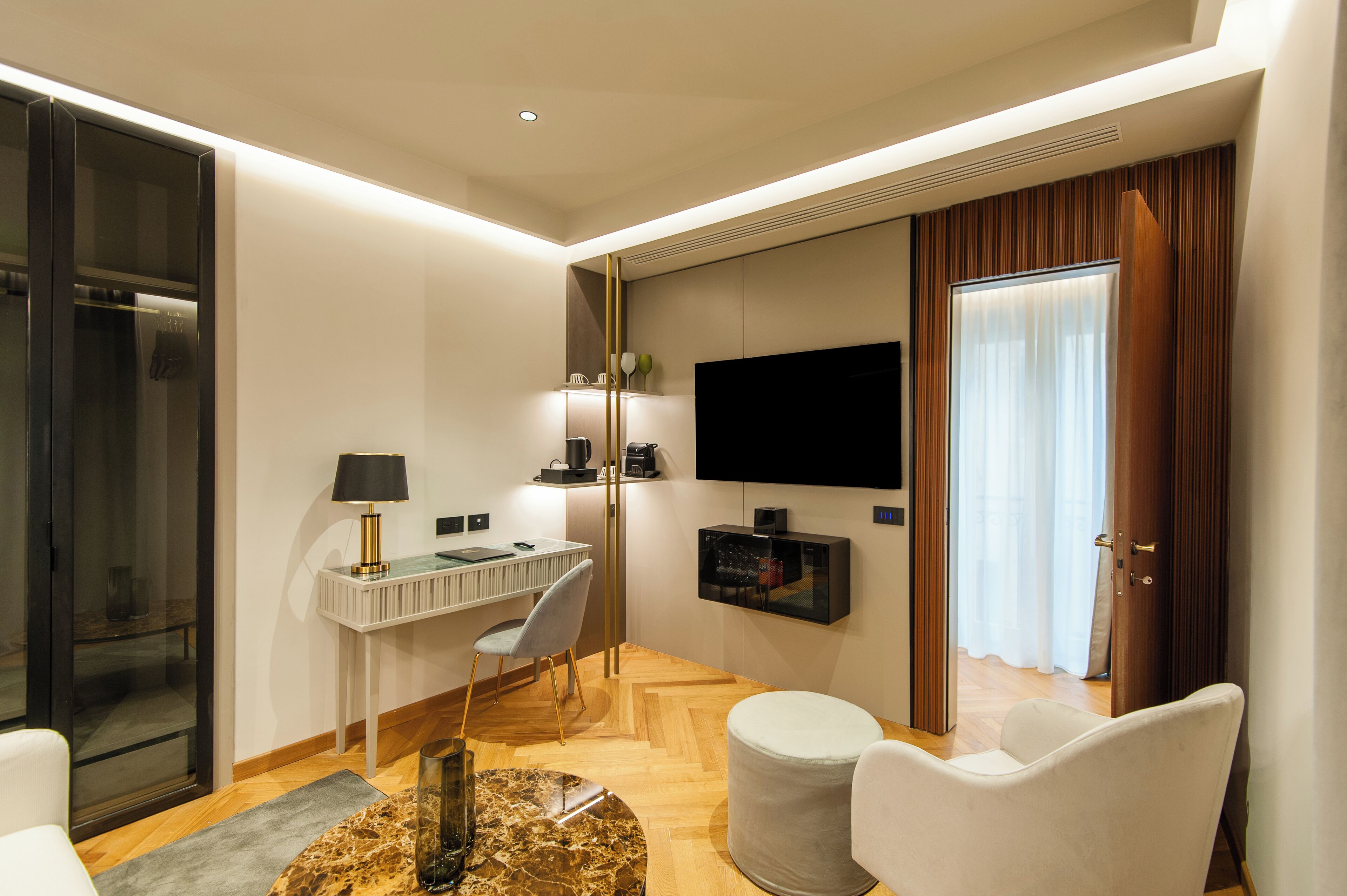 Quarto de hotel moderno com piso de madeira, uma escrivaninha com cadeira, televisão de tela plana na parede e uma janela grande com cortinas.