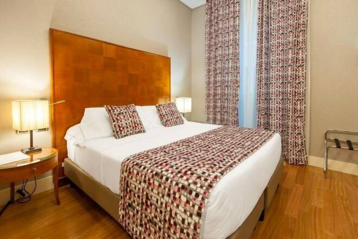 Habitación de hotel con cabecera de madera, cama con ropa de cama estampada, dos lámparas de noche y cortinas estampadas.
