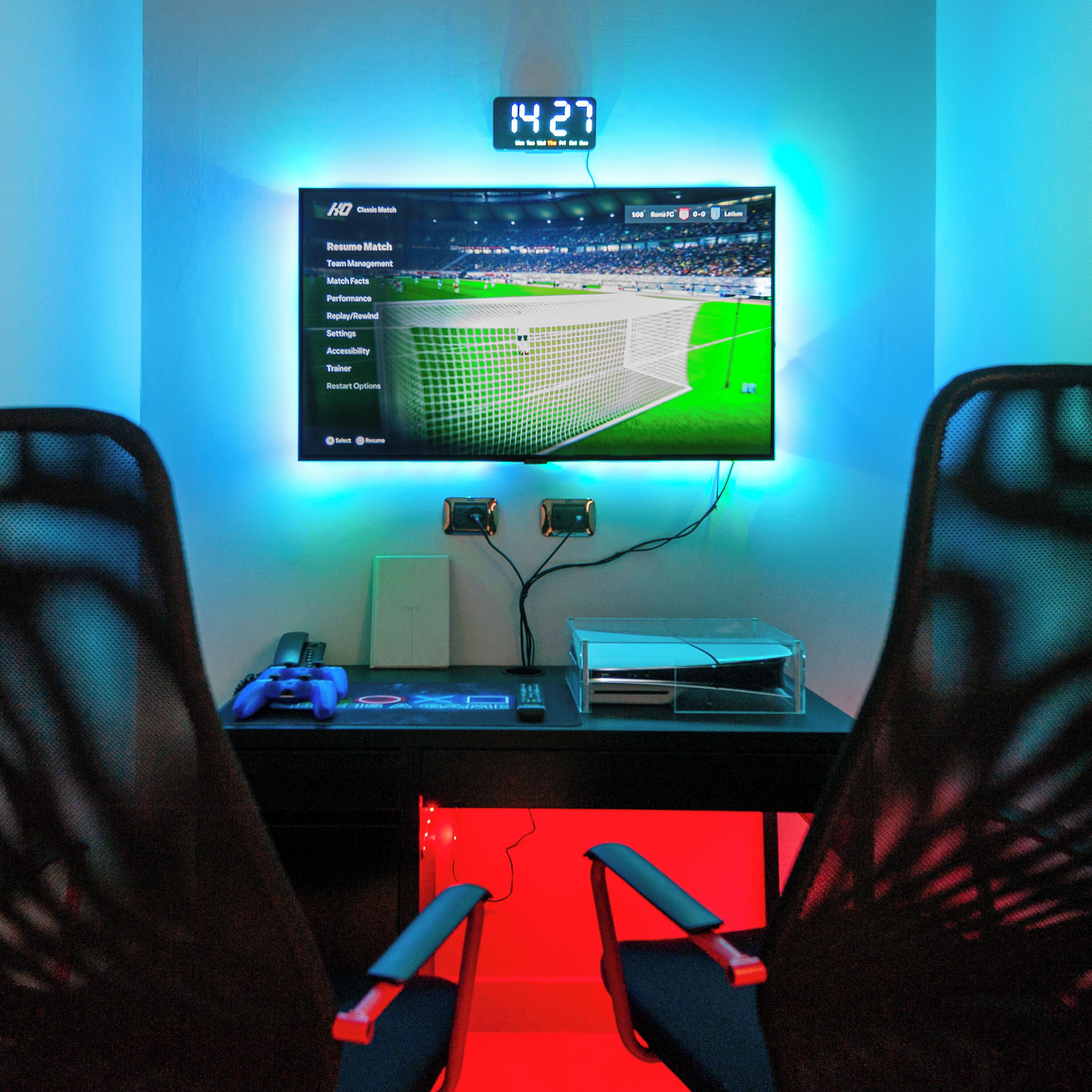 En gaming-opsætning med en skærm, der viser en fodboldkamp, to gaming-stole, et skrivebord med tastatur, mus og en game controller.