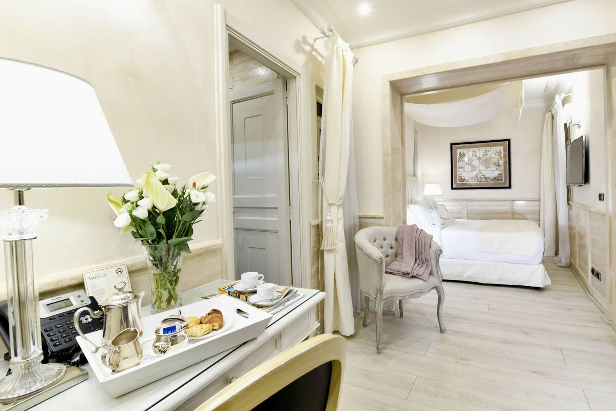 A hotel room with a bed, a bedside table with a lamp, a chair, a painting on the wall, and a bathroom visible through an open door.