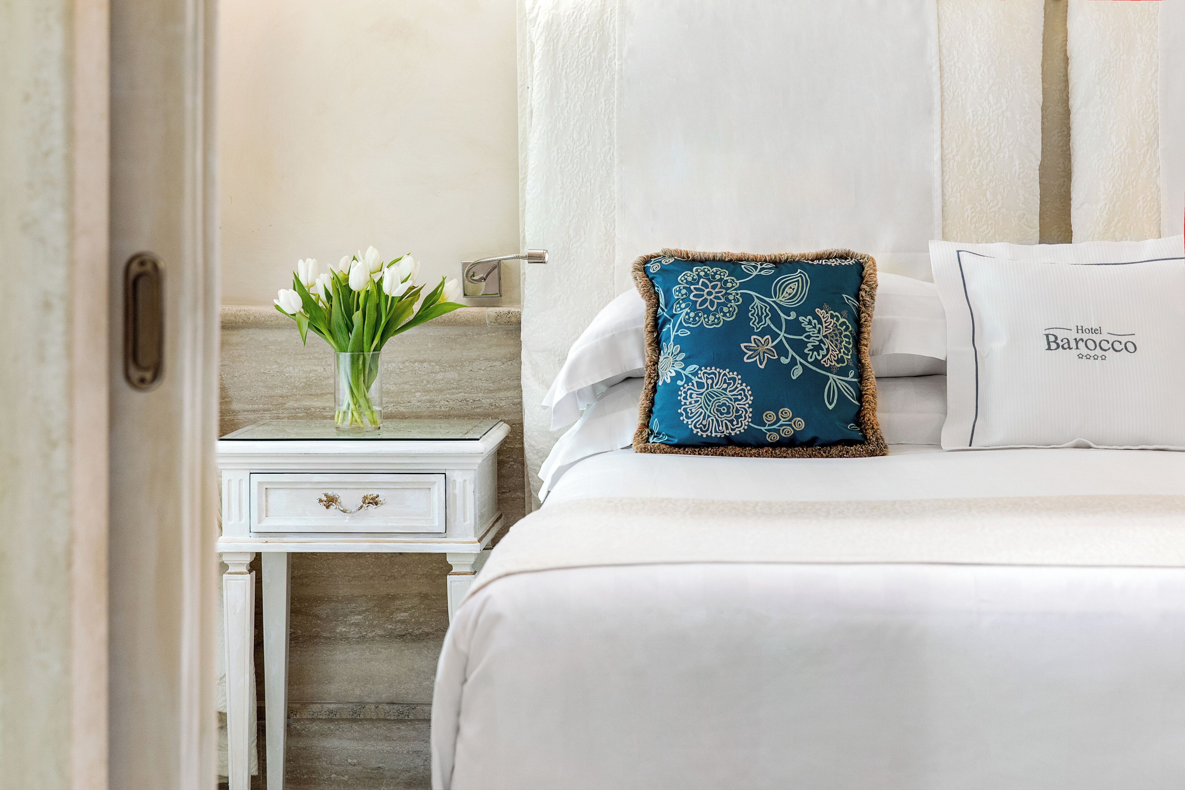 A hotel room with a bed, a nightstand, a vase with flowers, and a pillow with a floral pattern.