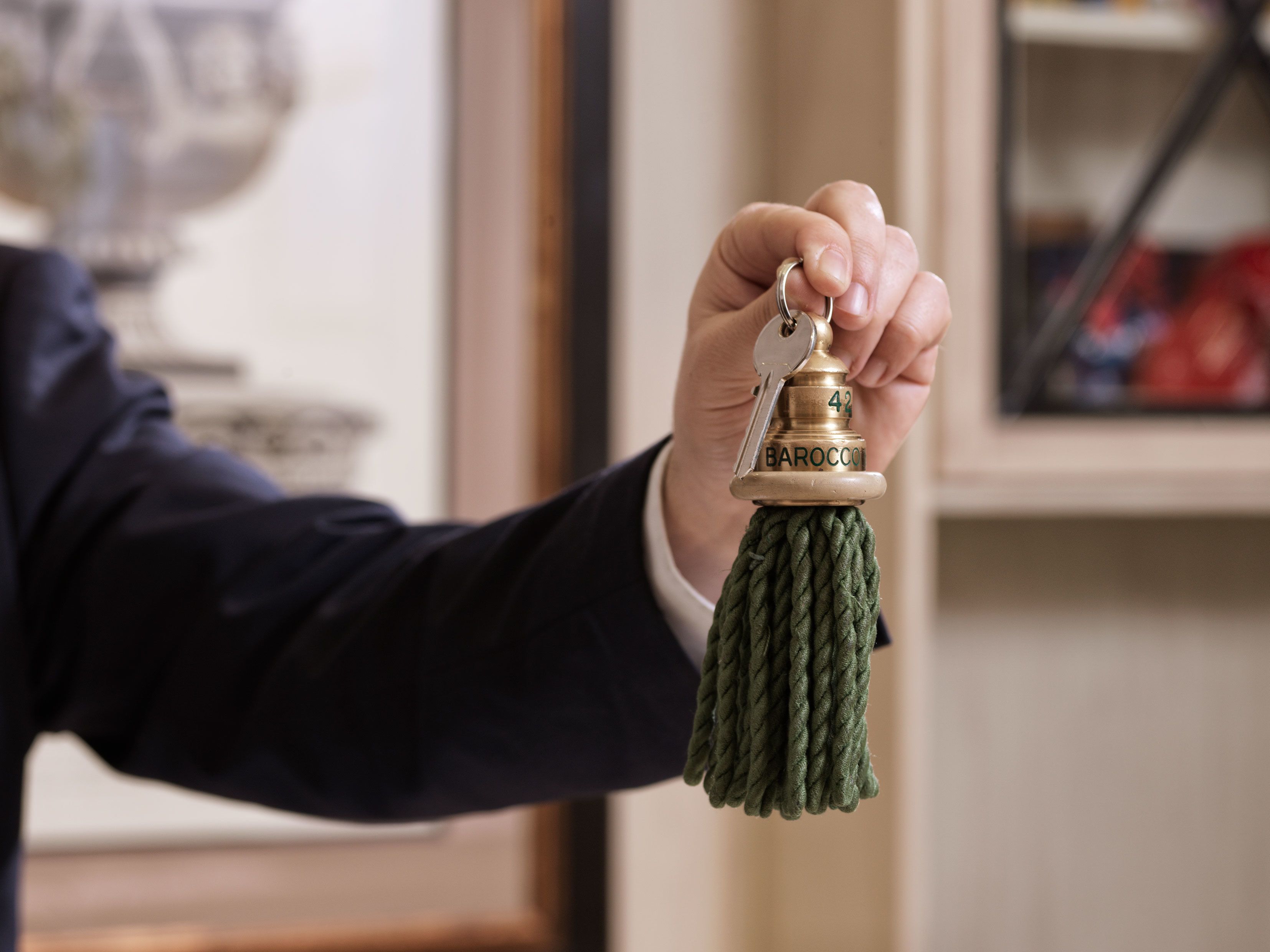 A hand holding a brass key with a green tassel, with a blurred background including a framed picture and a vase.