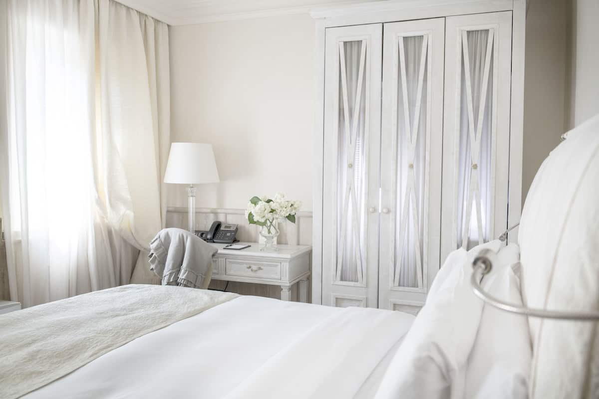 A hotel room with a bed, a bedside table with a lamp, a phone, and a vase with flowers. There is a wardrobe with curtains.