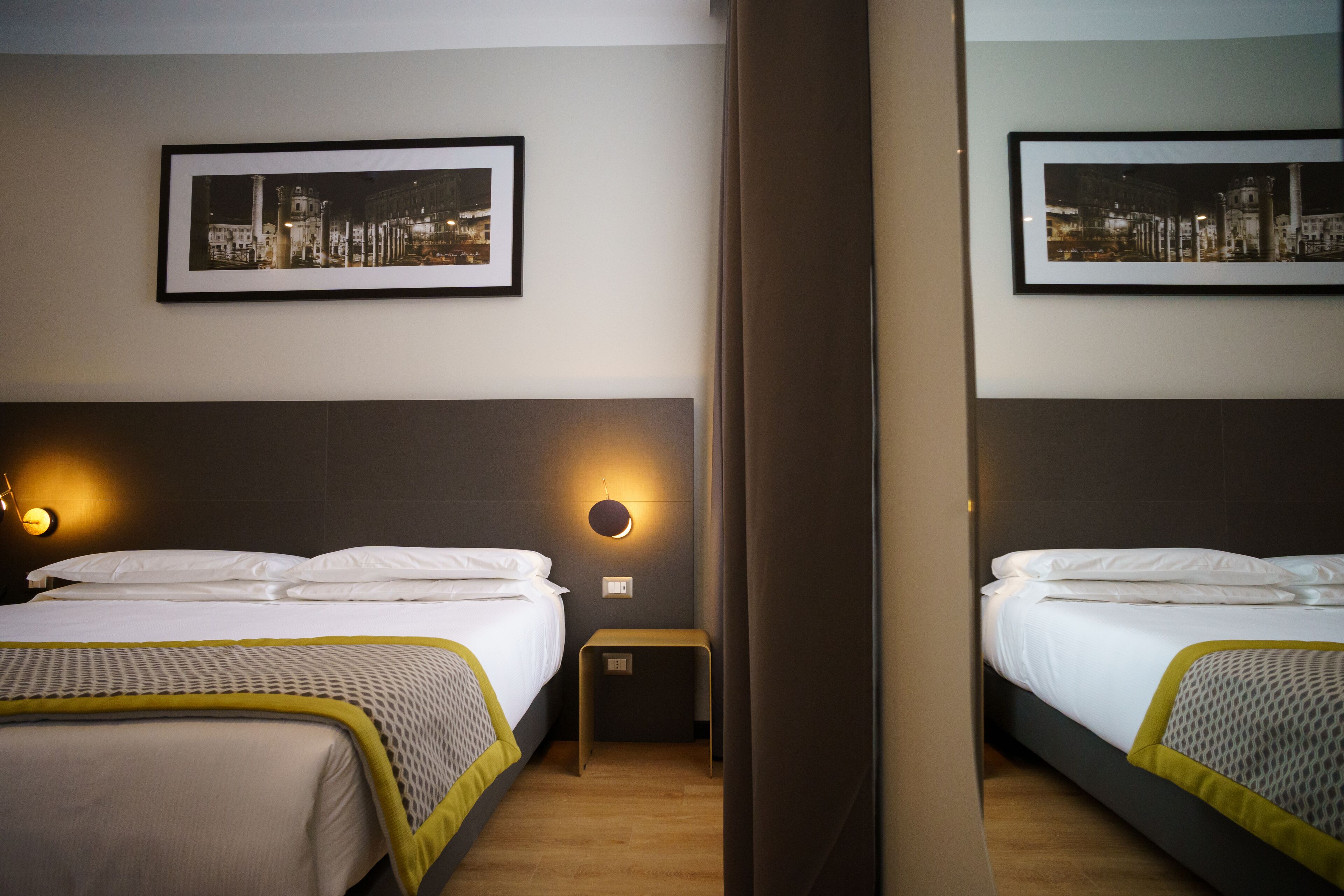 A hotel room with two beds, a nightstand, a wall-mounted lamp, and two framed pictures.