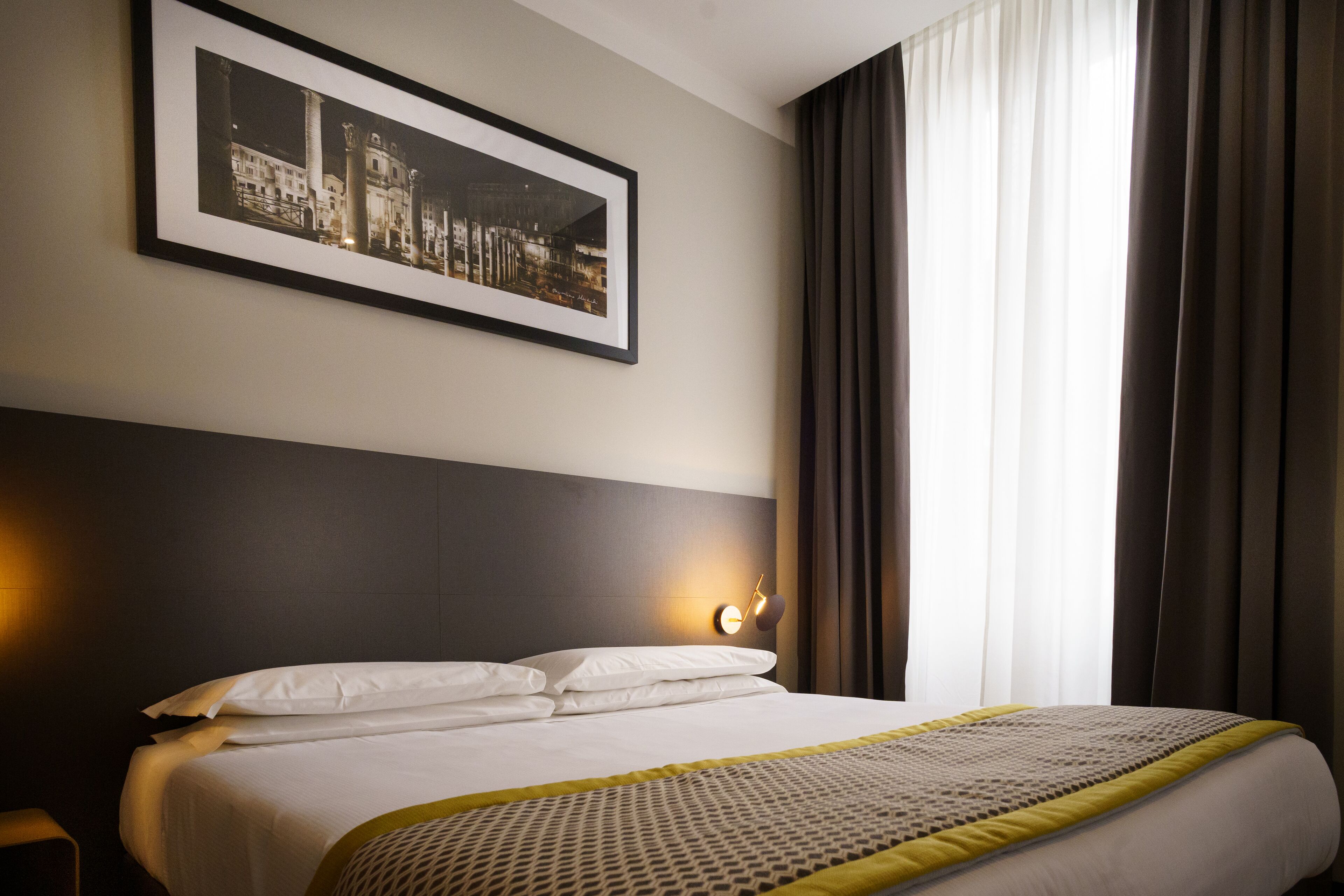 A neatly made bed with white linens and a patterned throw, a framed picture above the bed, and a window with curtains.