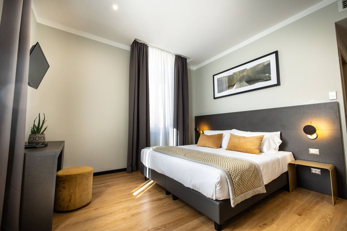 A modern hotel room with a large bed, a bedside table, a small stool, and a wall-mounted TV.
