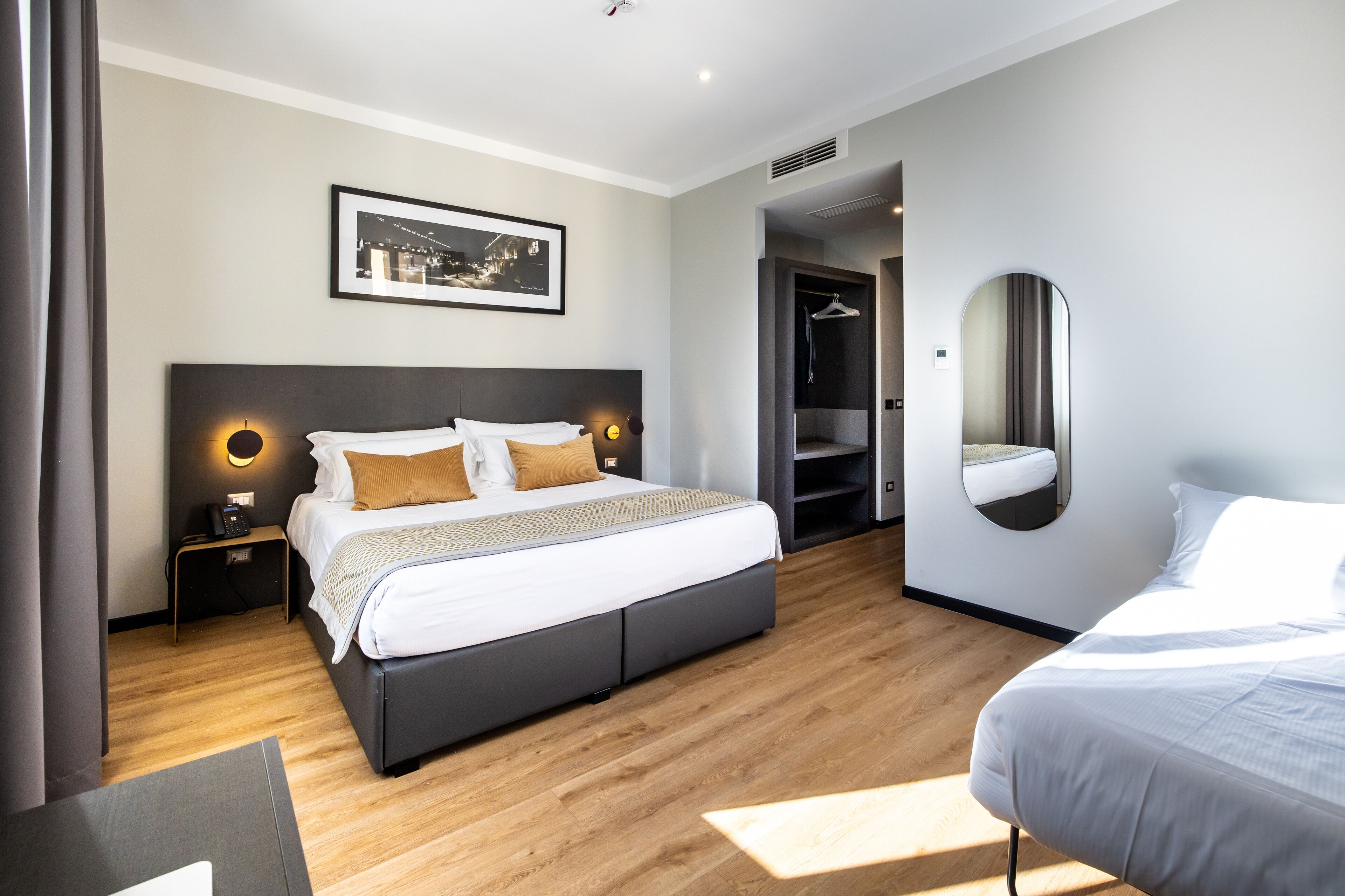 A modern hotel room with a large bed, bedside tables with lamps, a walk-in closet, and a large mirror.