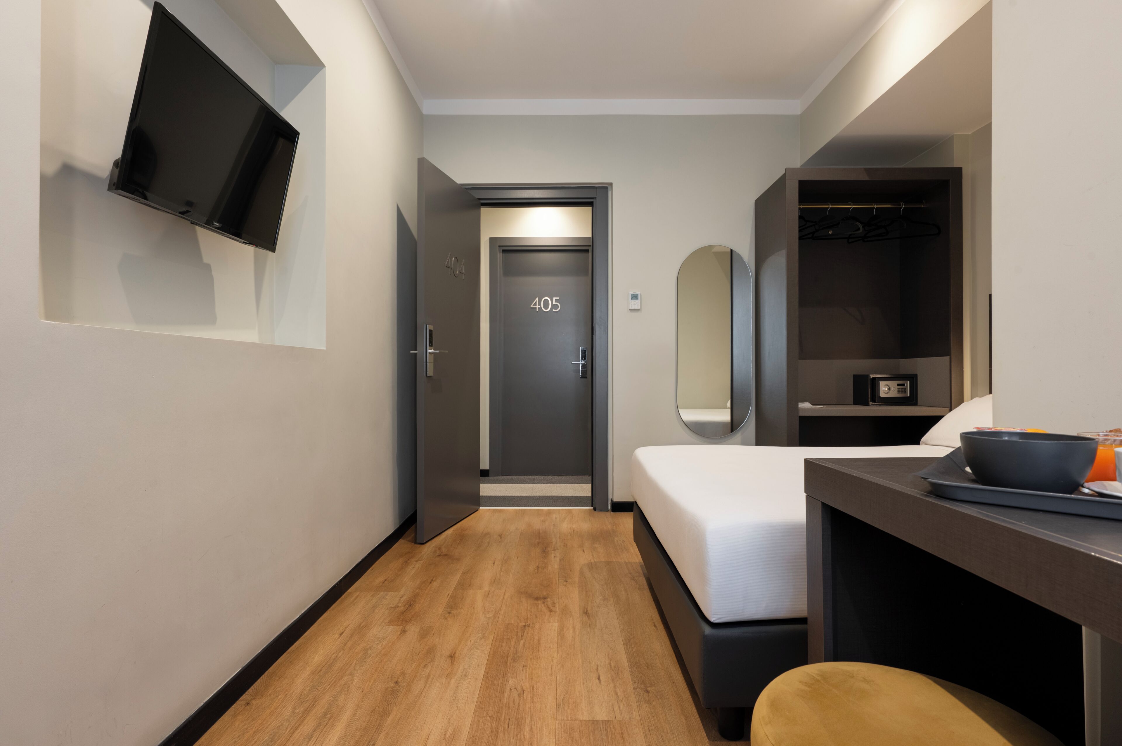 A modern hotel room with a bed, a desk with a bowl and orange drink, a TV mounted on the wall, and a door marked with the number 405.
