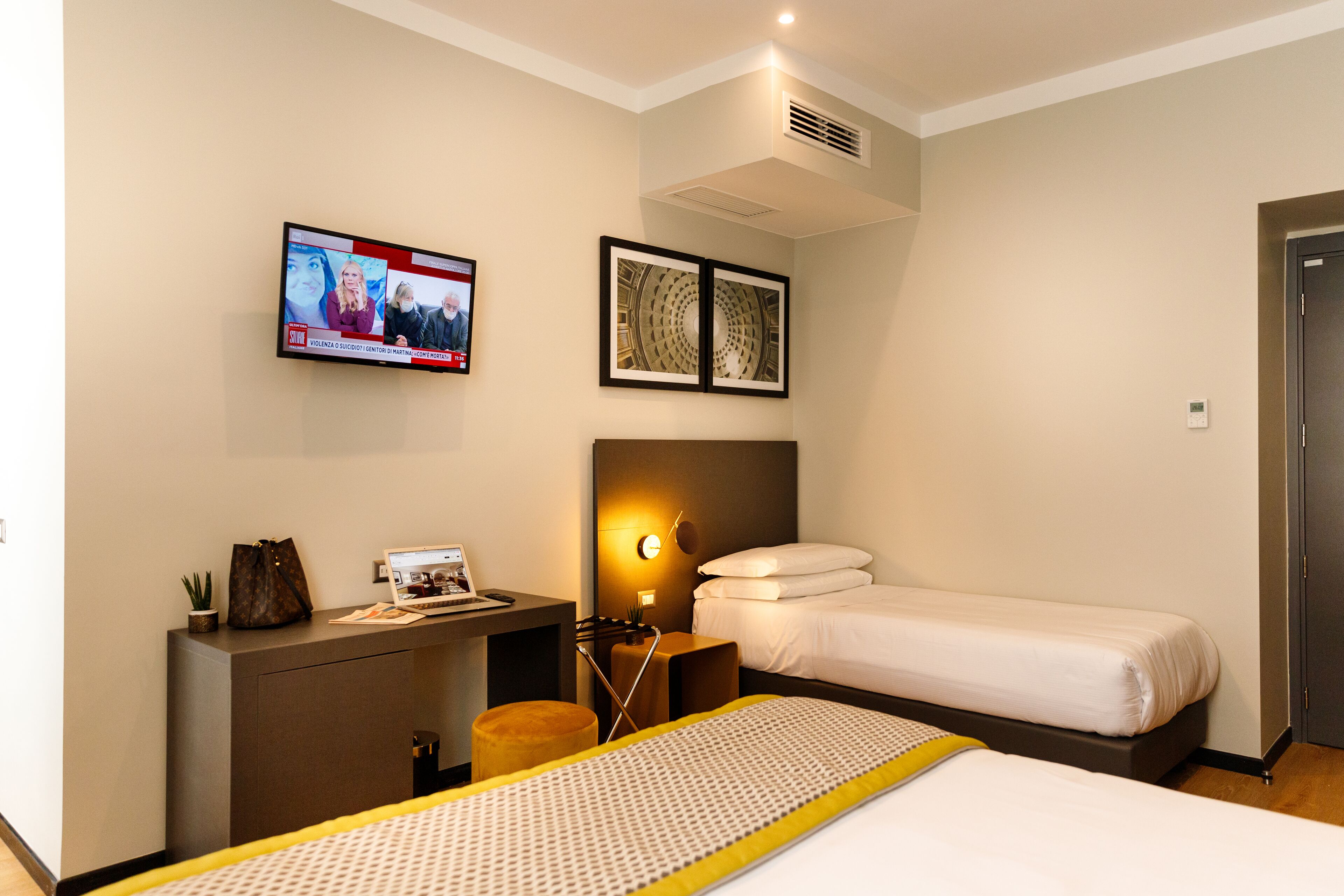 A hotel room with a bed, a desk with a laptop, a TV mounted on the wall, and a framed picture above the bed.