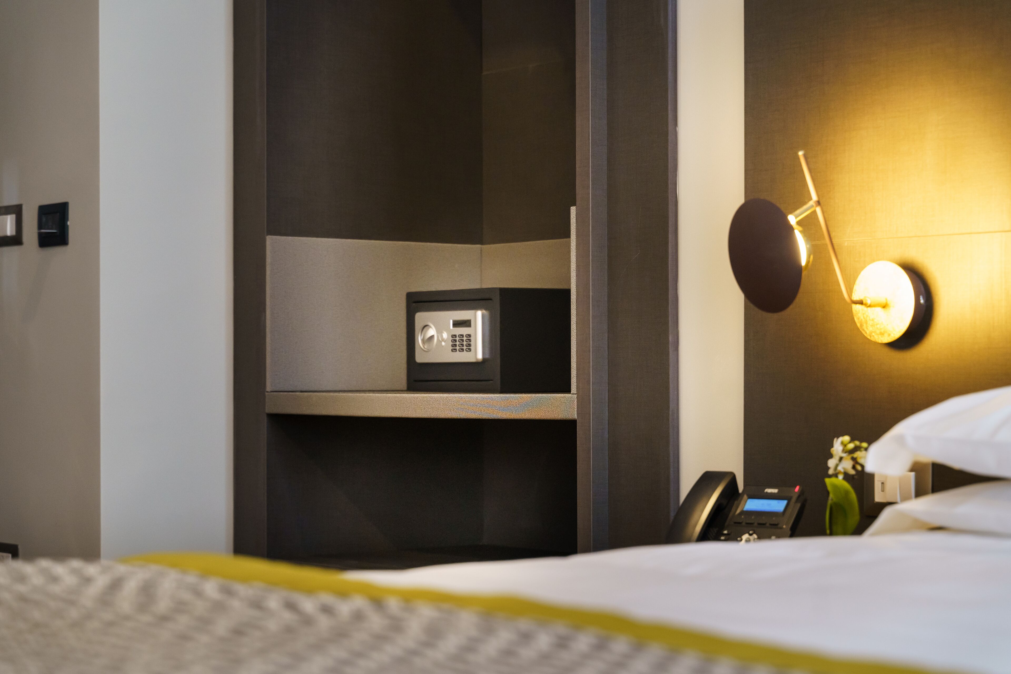 A hotel room with a bed, a bedside table with a phone, a small shelf with a safe, and a wall-mounted light fixture.