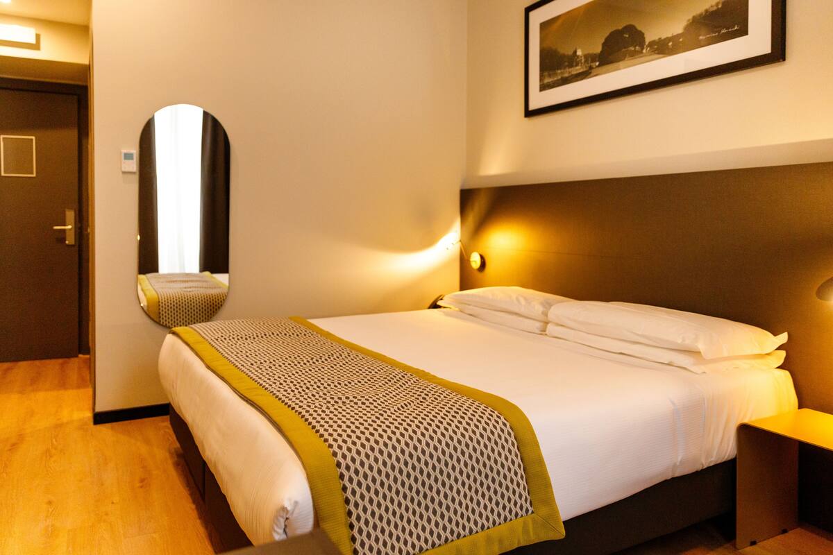 A neatly made bed with a patterned bedspread, a bedside table with a lamp, a wall-mounted mirror, and a framed picture above the bed.