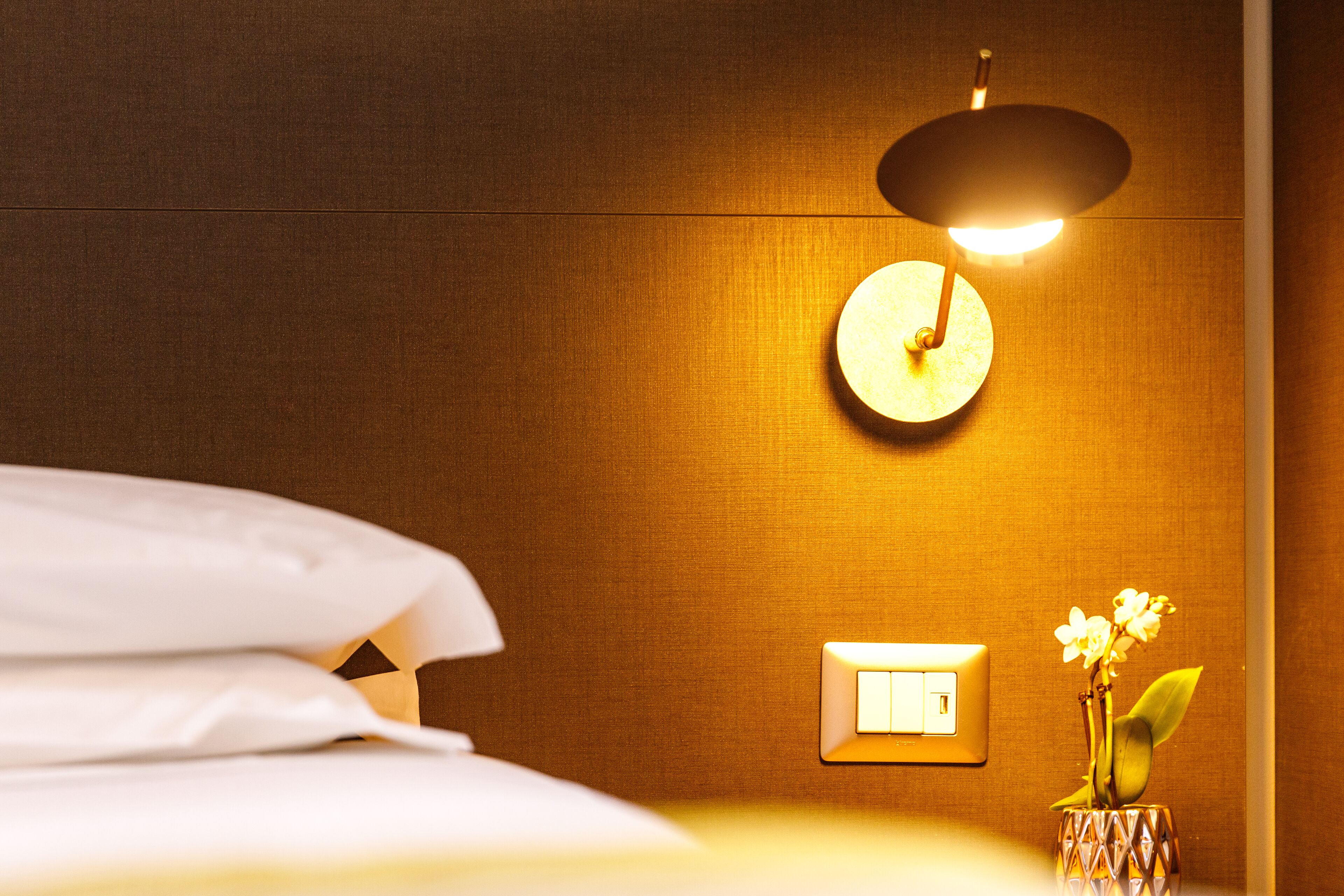 A hotel room with a bed, a wall-mounted light, a power outlet, and a vase with flowers.