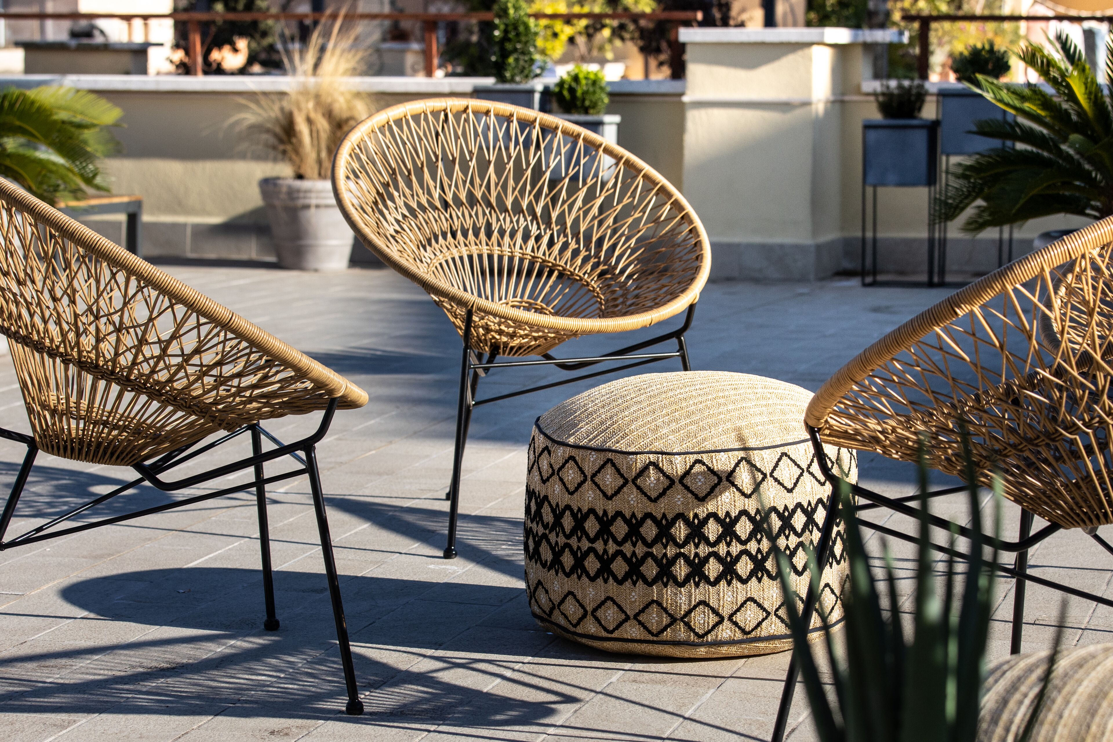 A rooftop terrace with wicker furniture and a round ottoman, surrounded by potted plants and a clear view of the city skyline.
