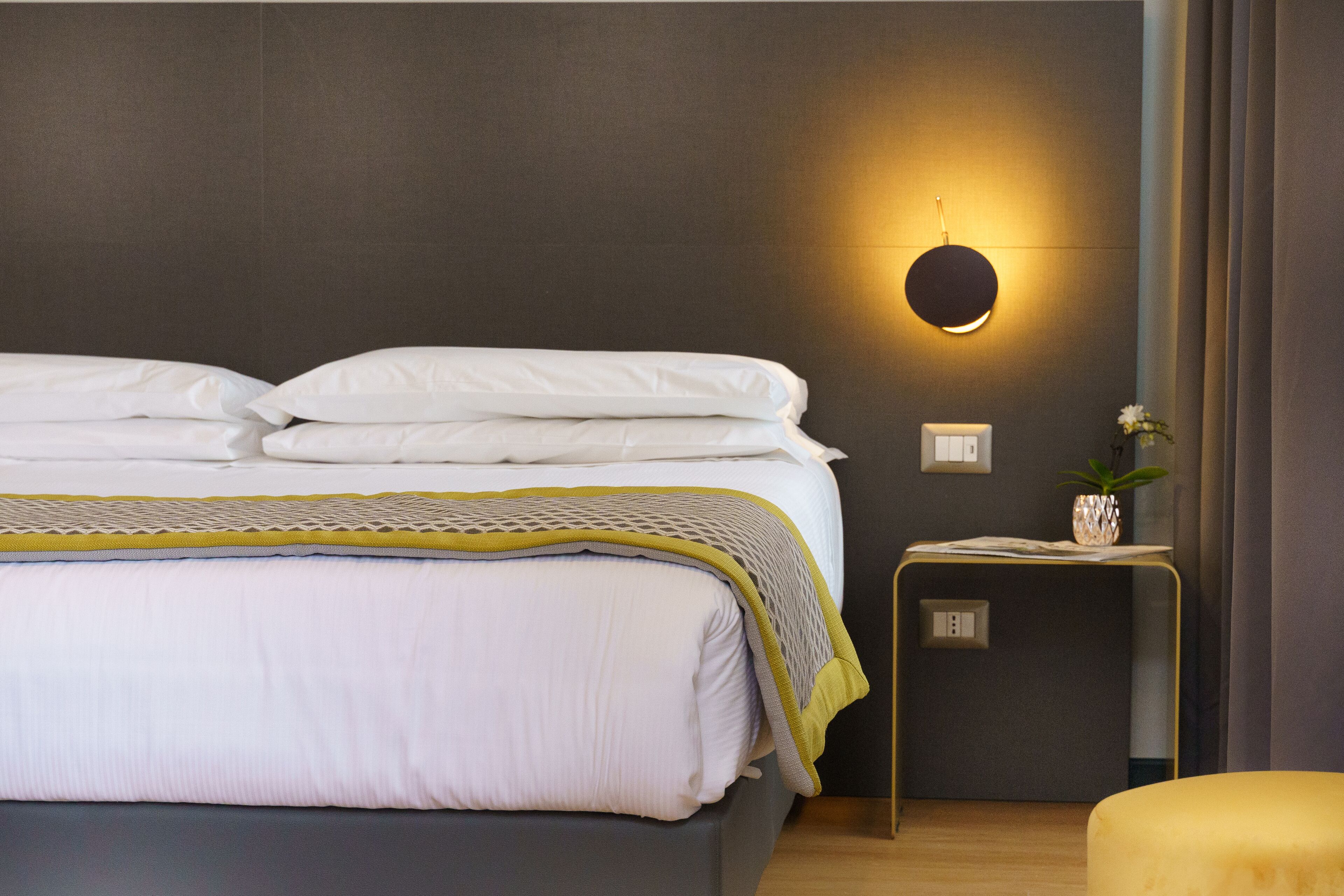 Una cama bien tendida con sábanas blancas y un detalle amarillo, una lámpara de pared, una mesita con una planta y suelo de madera.