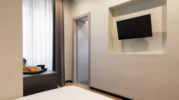Una camera d'albergo con un letto, una TV a schermo piatto fissata al muro, una finestra con tende e un vassoio con articoli per la colazione sul comodino.